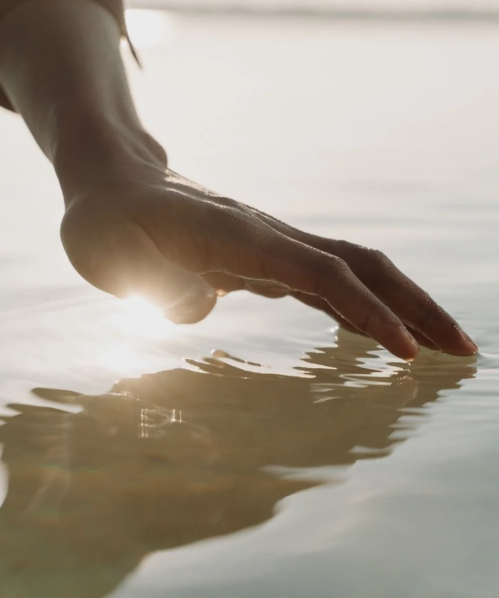 Close-up of a hand gently skimming the surface of calm water illuminated by soft sunlight, evoking mindfulness, somatic awareness, and nervous system regulation.