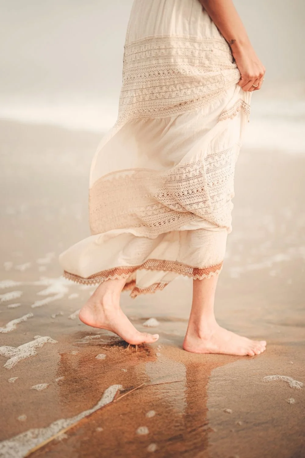 Barefoot person in a flowing white dress walking along the shoreline, capturing a sense of freedom, grounding, and connection with nature.