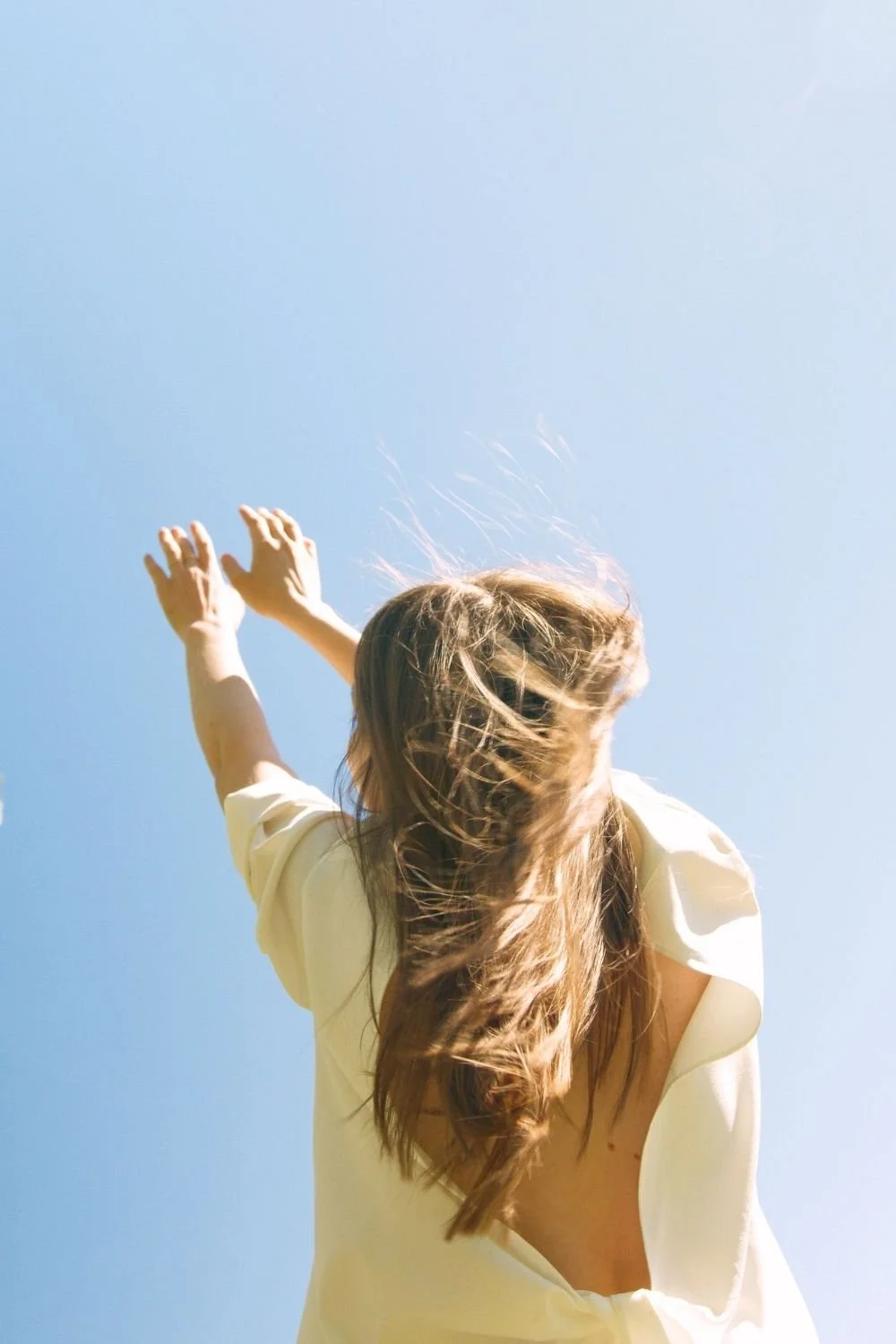 Person with windblown hair reaching both arms toward a clear blue sky, expressing freedom, experiential wellness, and connection to mind body health.