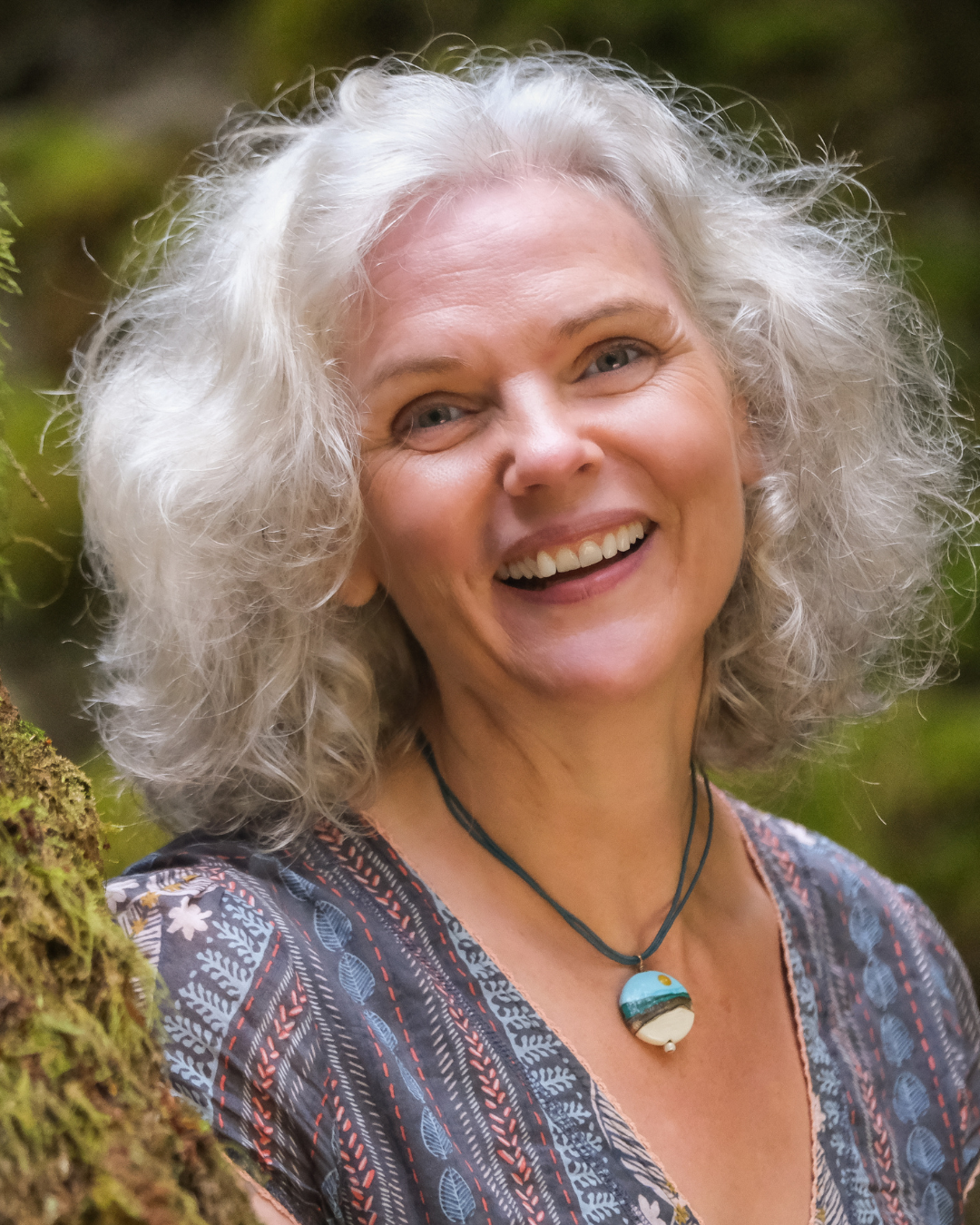A smiling elderly woman with curly gray hair wearing a patterned blouse and a necklace with a large round pendant, standing outdoors near a moss-covered tree with green foliage in the background.