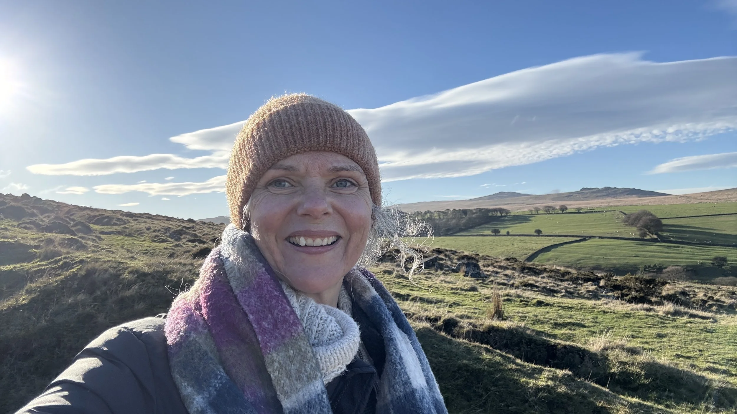 A smiling woman wearing a brown knit hat and multicolored scarf standing outdoors in a rural landscape with rolling green hills, fenced pastures, and a blue sky with clouds.
