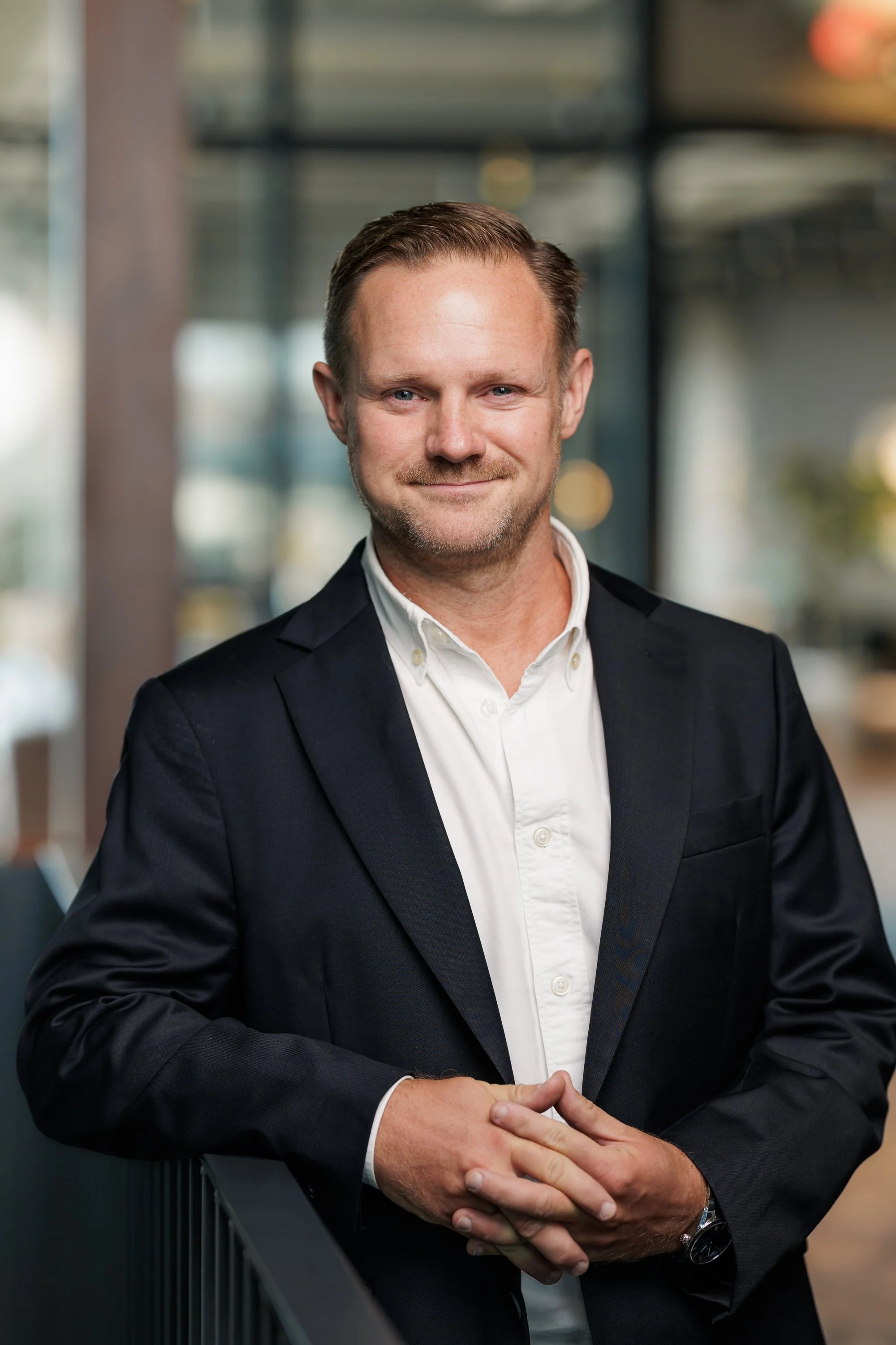 A professional man in a dark suit smiling, standing with hands clasped, in front of a modern office background with glass walls.