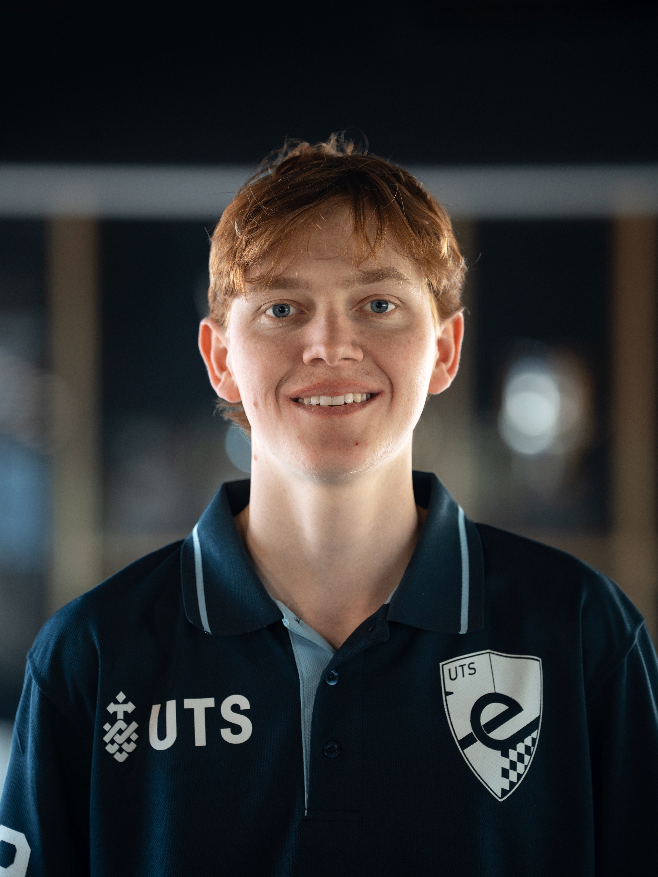 A young man with red hair and blue eyes wearing a navy blue sports jersey with logos and a crest, smiling and standing indoors with a blurred background.