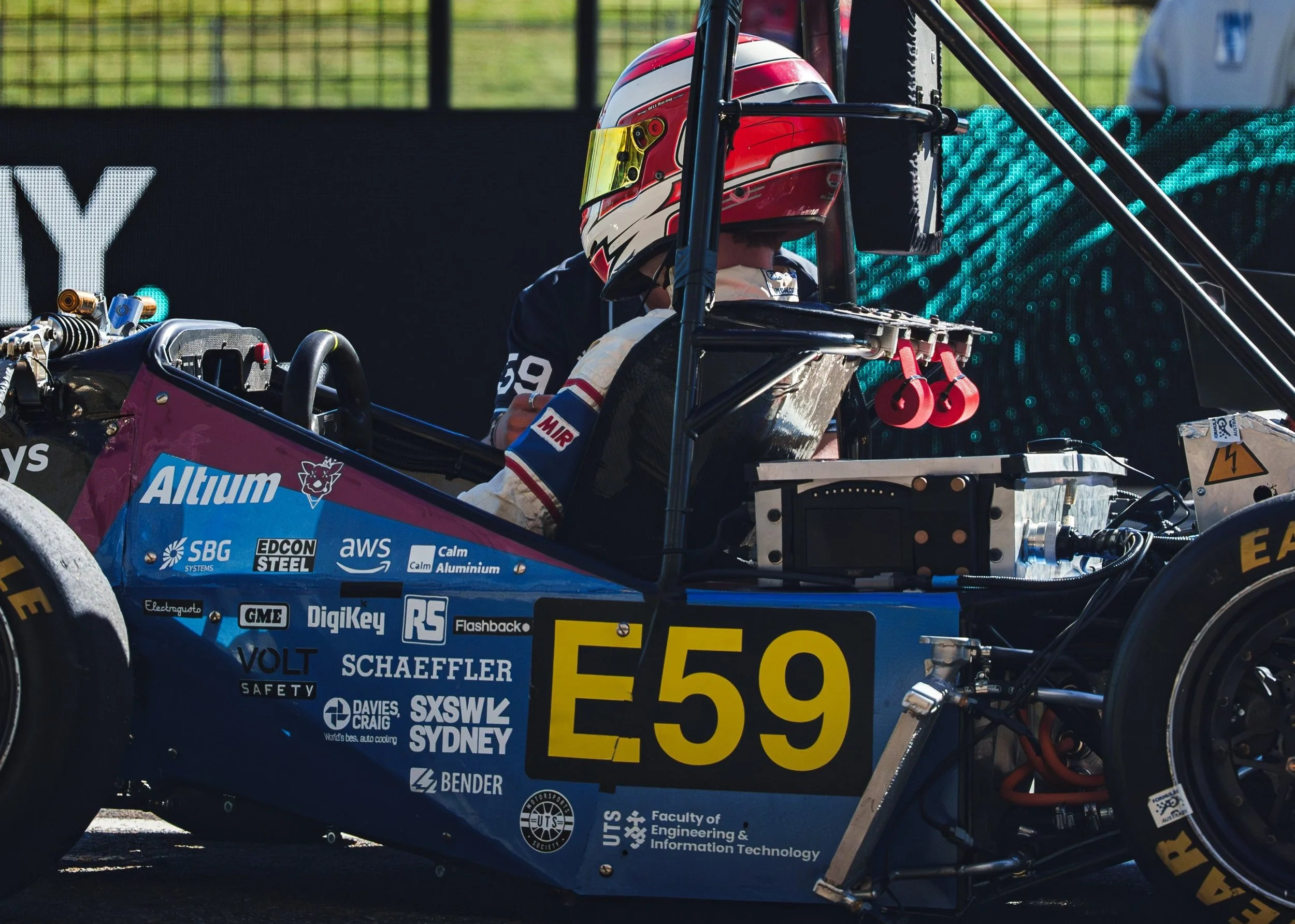 A race car driver wearing a red, white, and black helmet sitting inside a race car with various sponsor stickers. The car's body features logos and text, including 'Altium,' 'Schaeffer,' 'Syswix,' and 'UTS.'