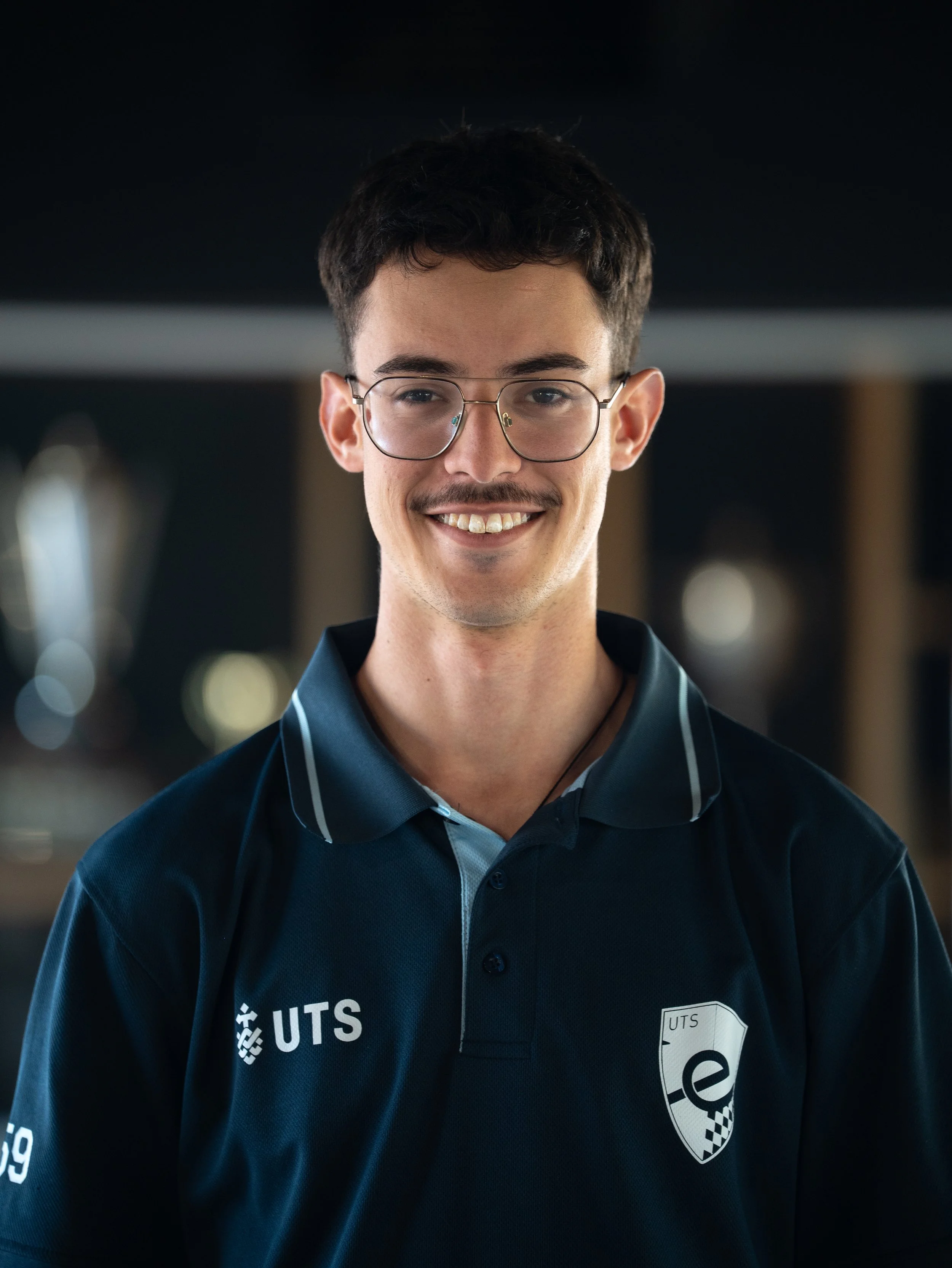 Young man wearing glasses and a dark blue UTS sports jersey, smiling, with a blurred indoor background.