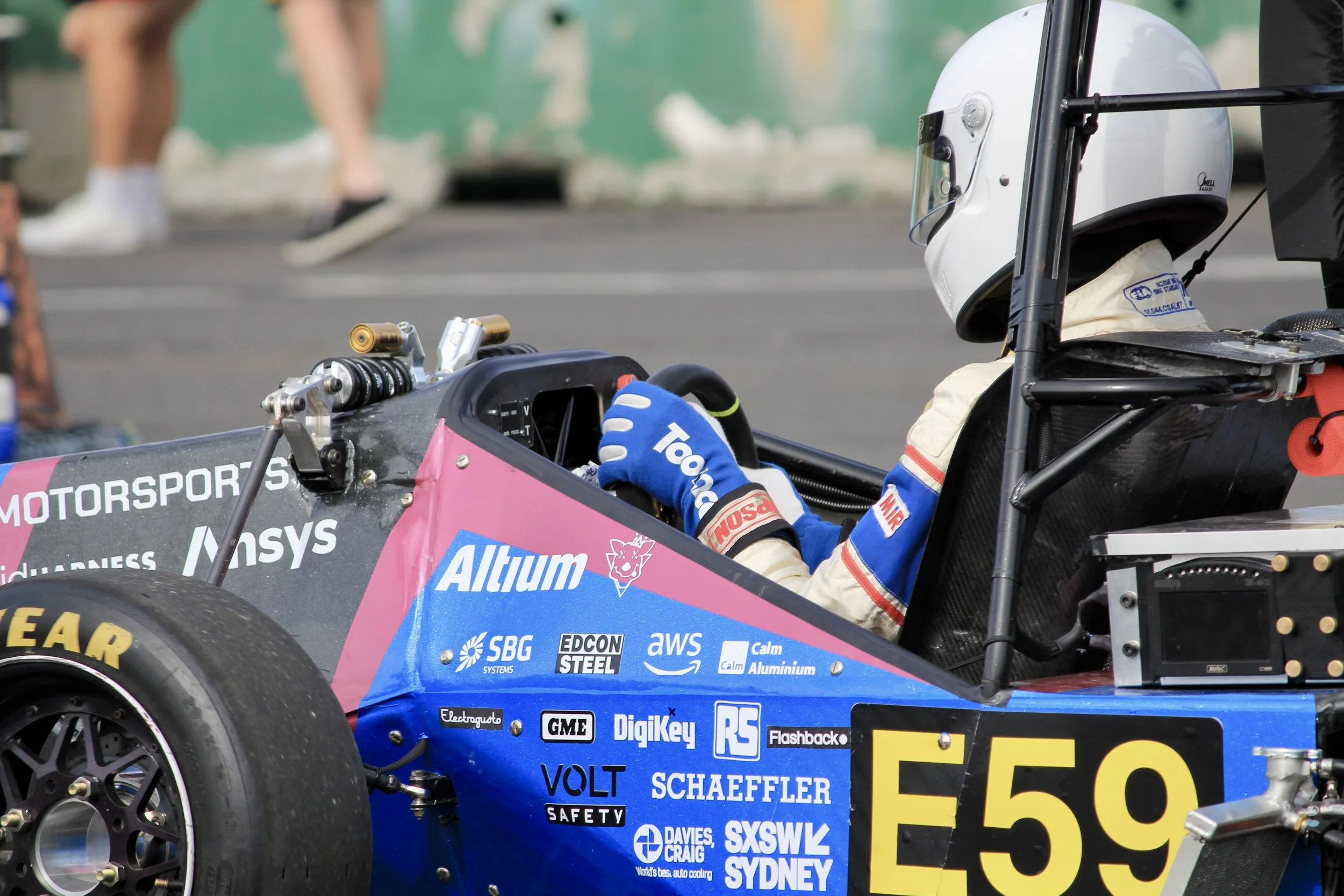 Race car driver wearing a helmet and racing suit sitting in a race car with sponsor logos, preparing for a race at a racing event.