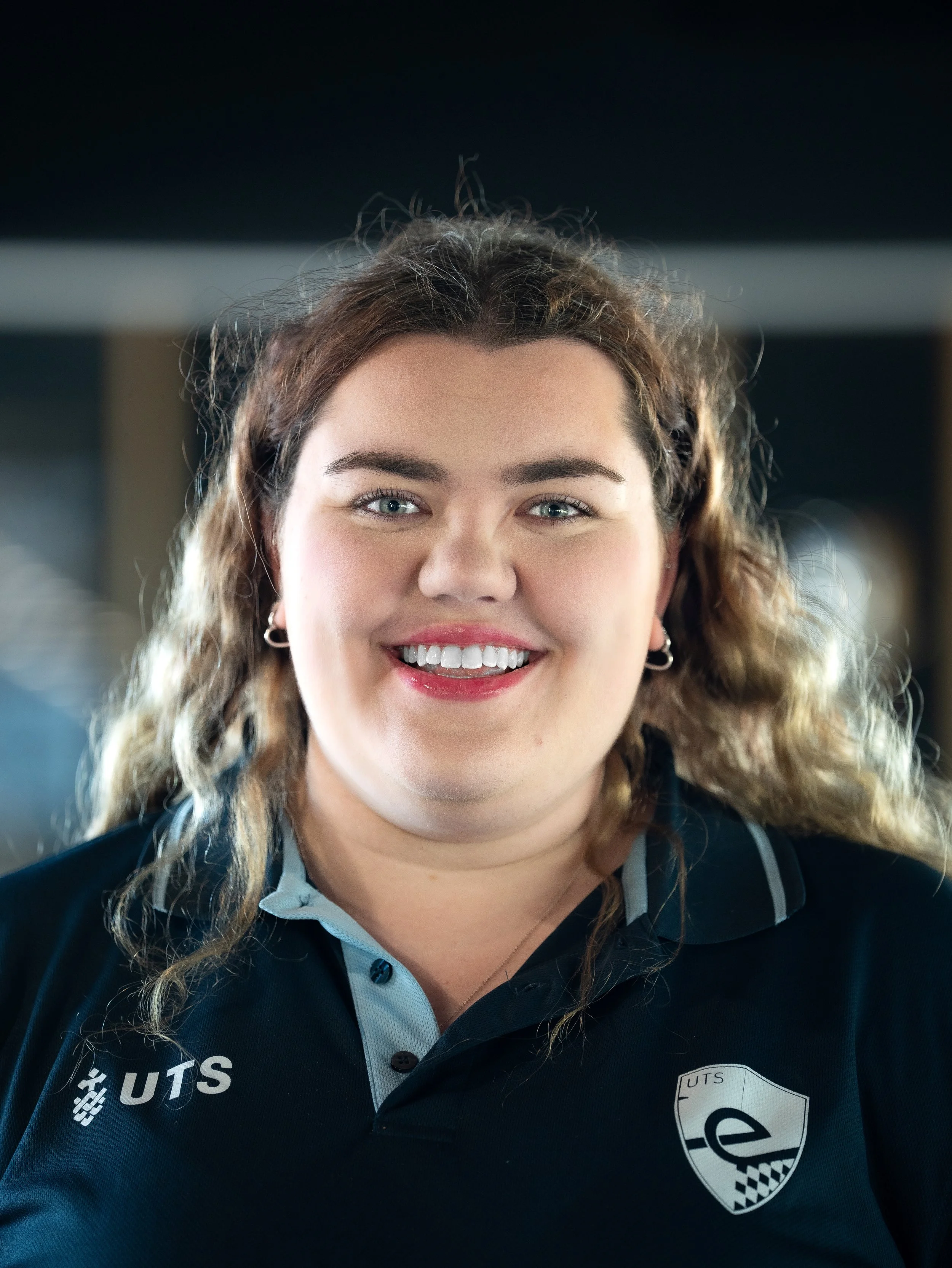 Close-up of a young woman with curly hair and blue eyes, smiling, wearing a black sports shirt with 'UTS' and a logo, indoors with blurred background.