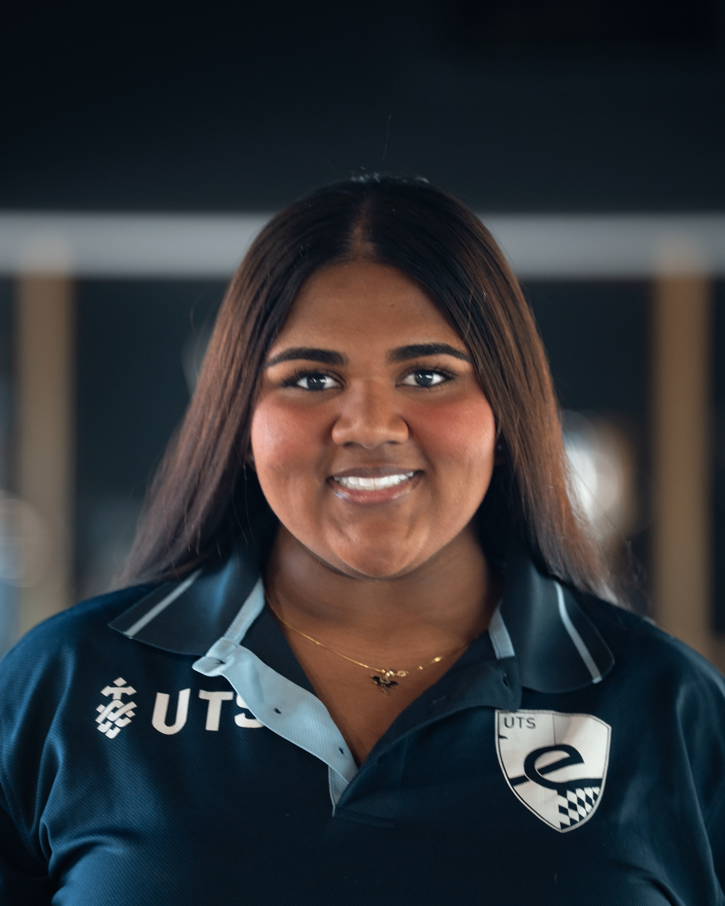 Close-up of a woman with brown skin and long brown hair, smiling, wearing a navy blue sports shirt with a logo that reads "UTS" and a small gold necklace, indoors with a blurred background.