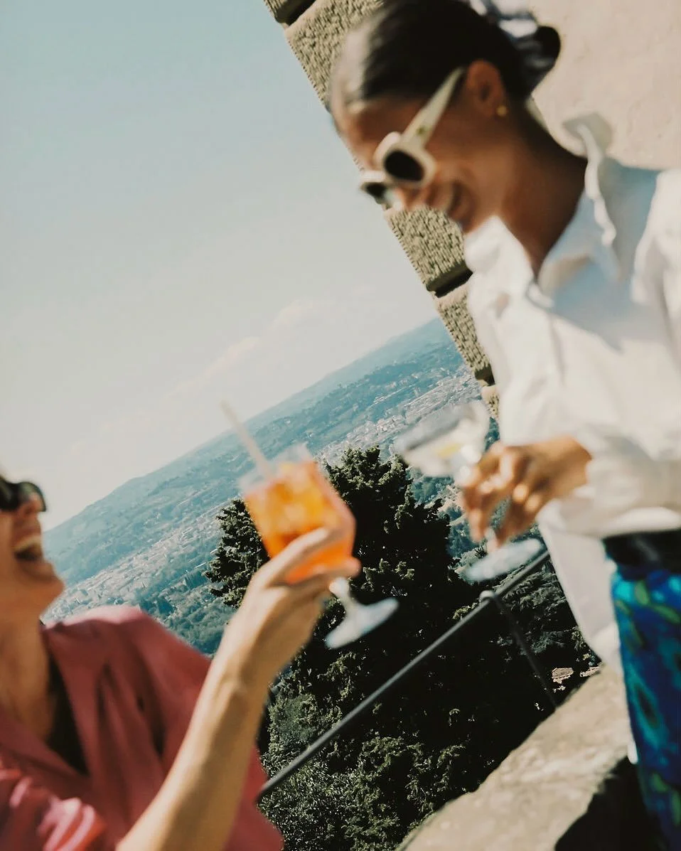 Two women enjoying drinks outdoors, with one woman in a white shirt and sunglasses and the other in a pink shirt, on a sunny day with scenic landscape and trees in the background.