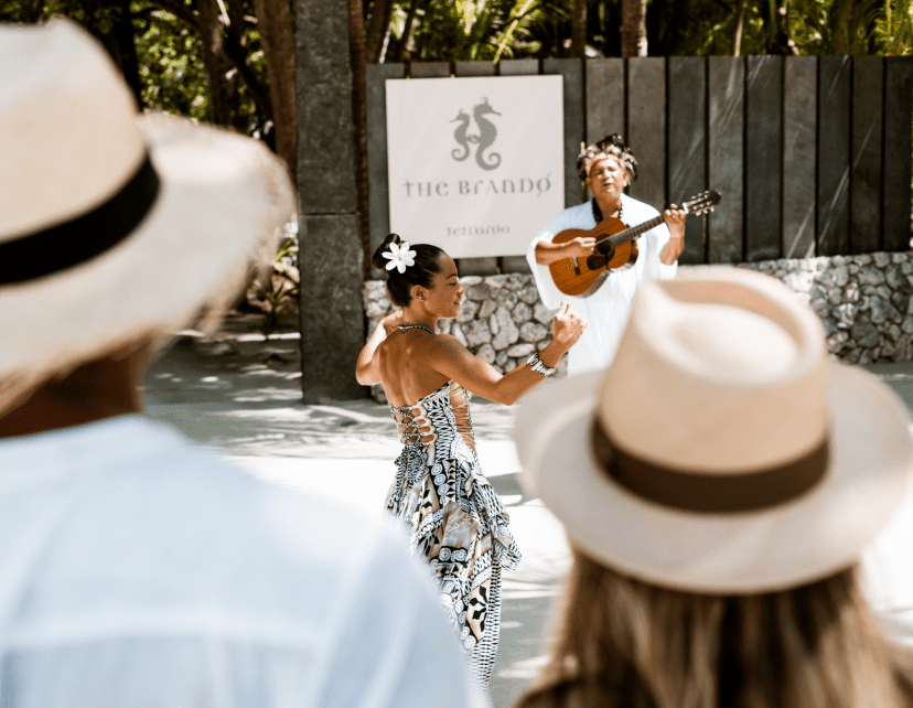 A woman in a patterned dress dances while another woman plays guitar and sings at an outdoor event, with an audience wearing straw hats watching.