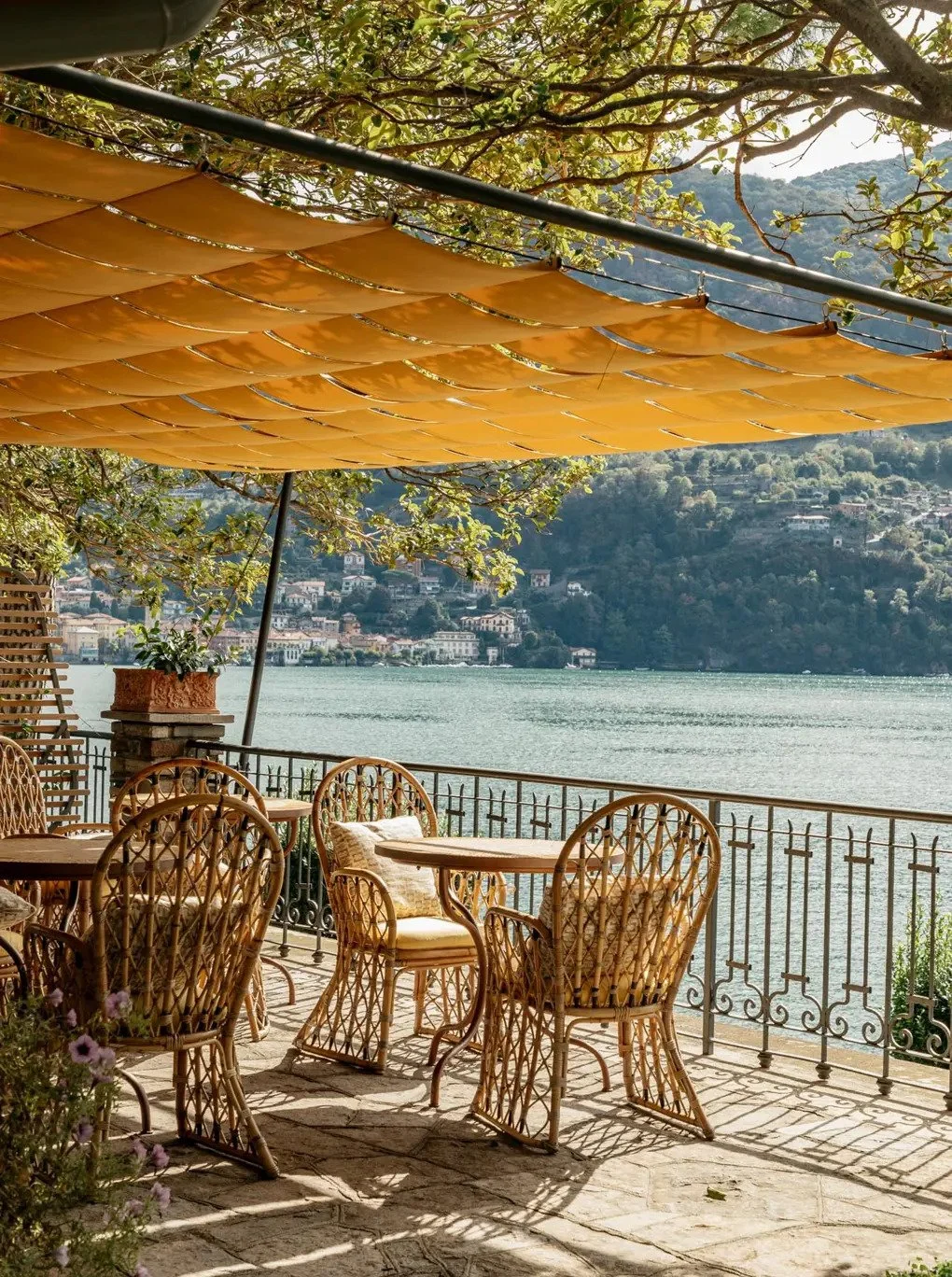 An outdoor terrace by a lake with wicker chairs and tables, shaded by a large yellow sunshade. In the background, there are green hills and a lakeside town.