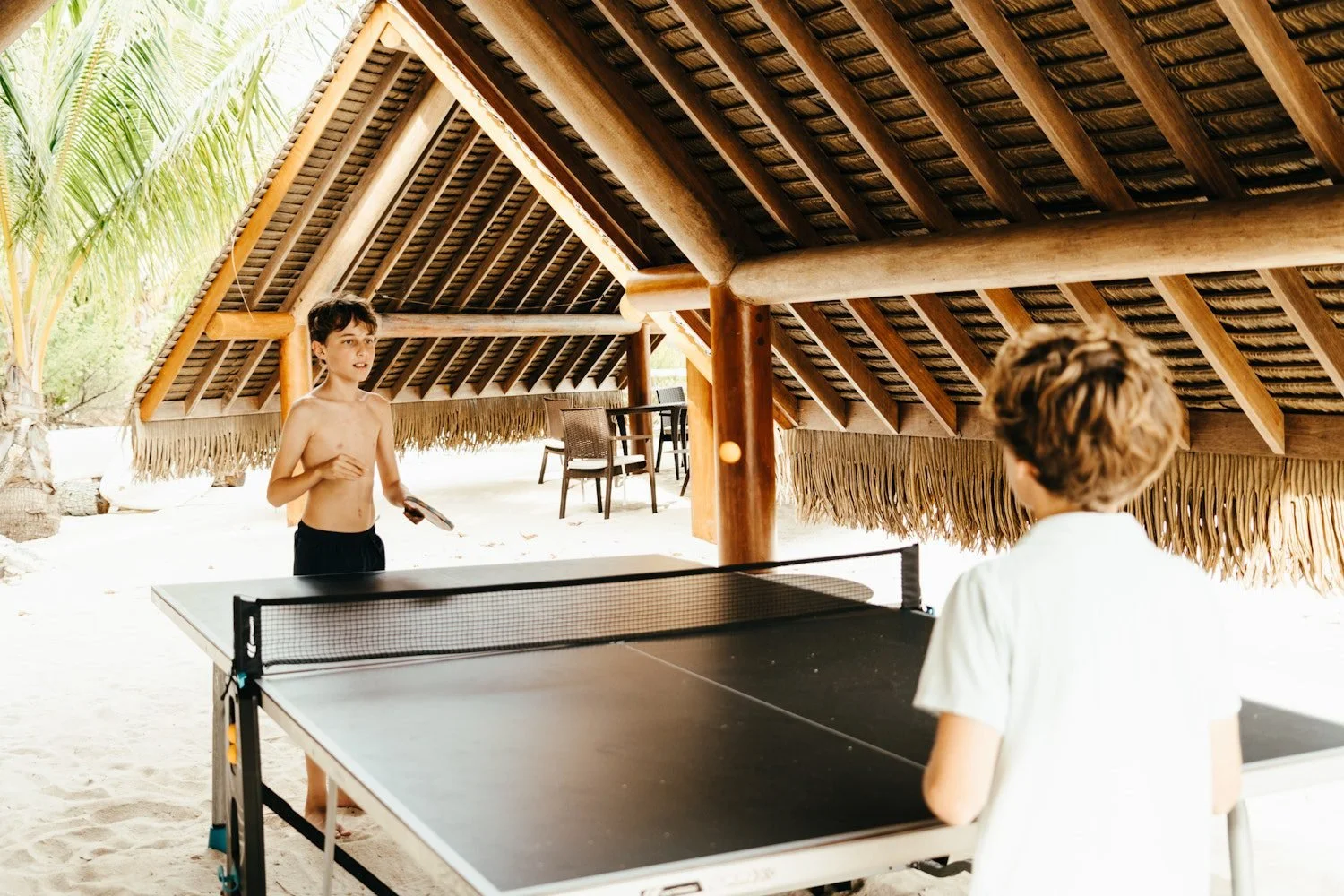 Two boys playing ping pong under a thatched roof hut on a beach, with chairs in the background and tropical trees nearby.