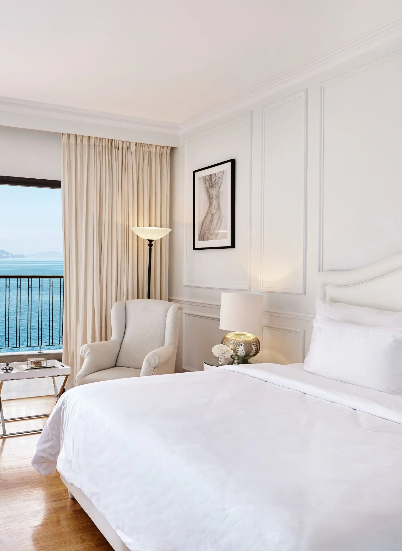 A hotel room with a view of the ocean, featuring a white bed with pillows, a beige armchair, a small side table with a flower arrangement, a table lamp, and a large window with beige curtains.