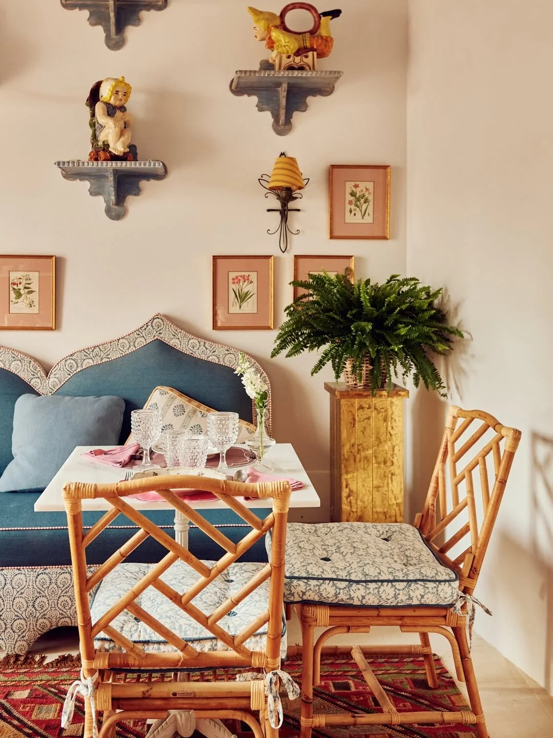 A cozy dining nook with a white table set with glassware and pink napkins, surrounded by bamboo chairs with cushions. In the background, a blue upholstered bench with a cushion, framed botanical prints on the wall, a potted fern on a wooden stand, and colorful decorative shelves with figurines and small objects.