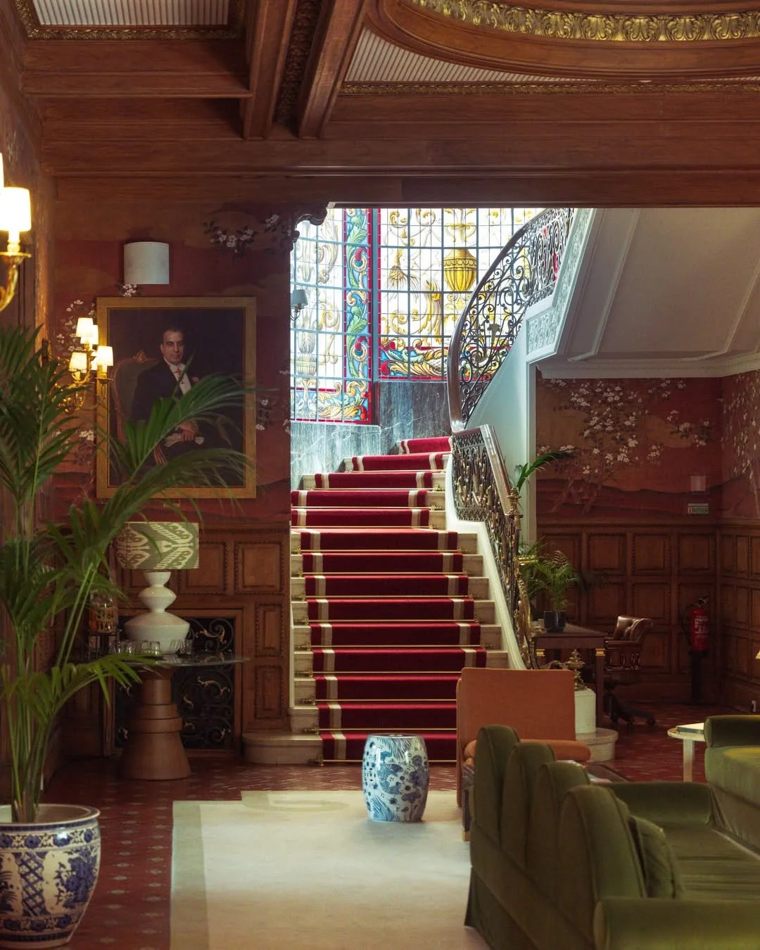Luxurious hotel interior featuring a grand staircase with red carpet, stained glass window at the top, framed portrait on the wall, plants, and elegant furniture.