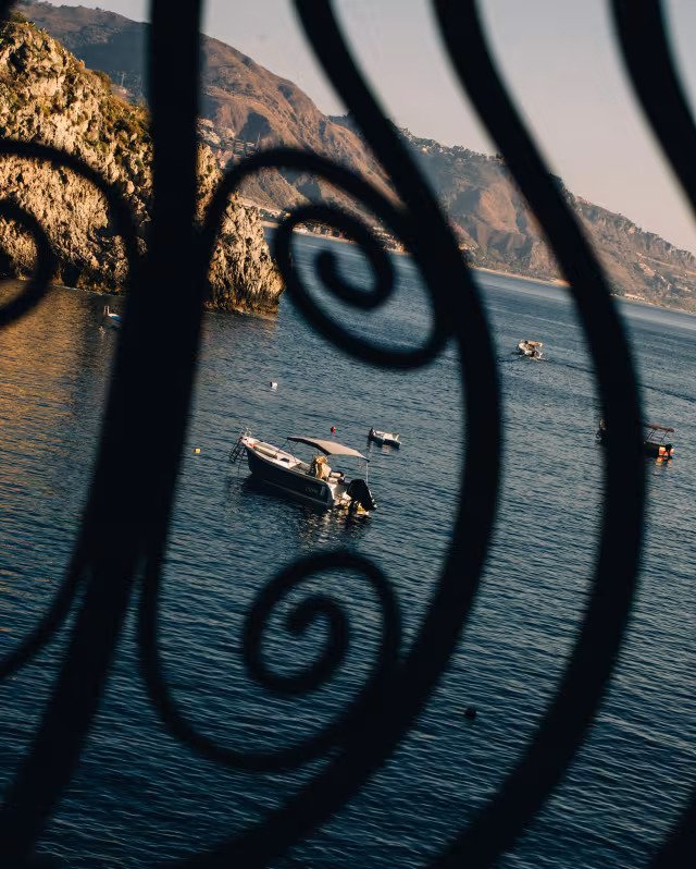 Boats anchored in a peaceful bay with rocky hills in the background, viewed through a decorative wrought iron railing.
