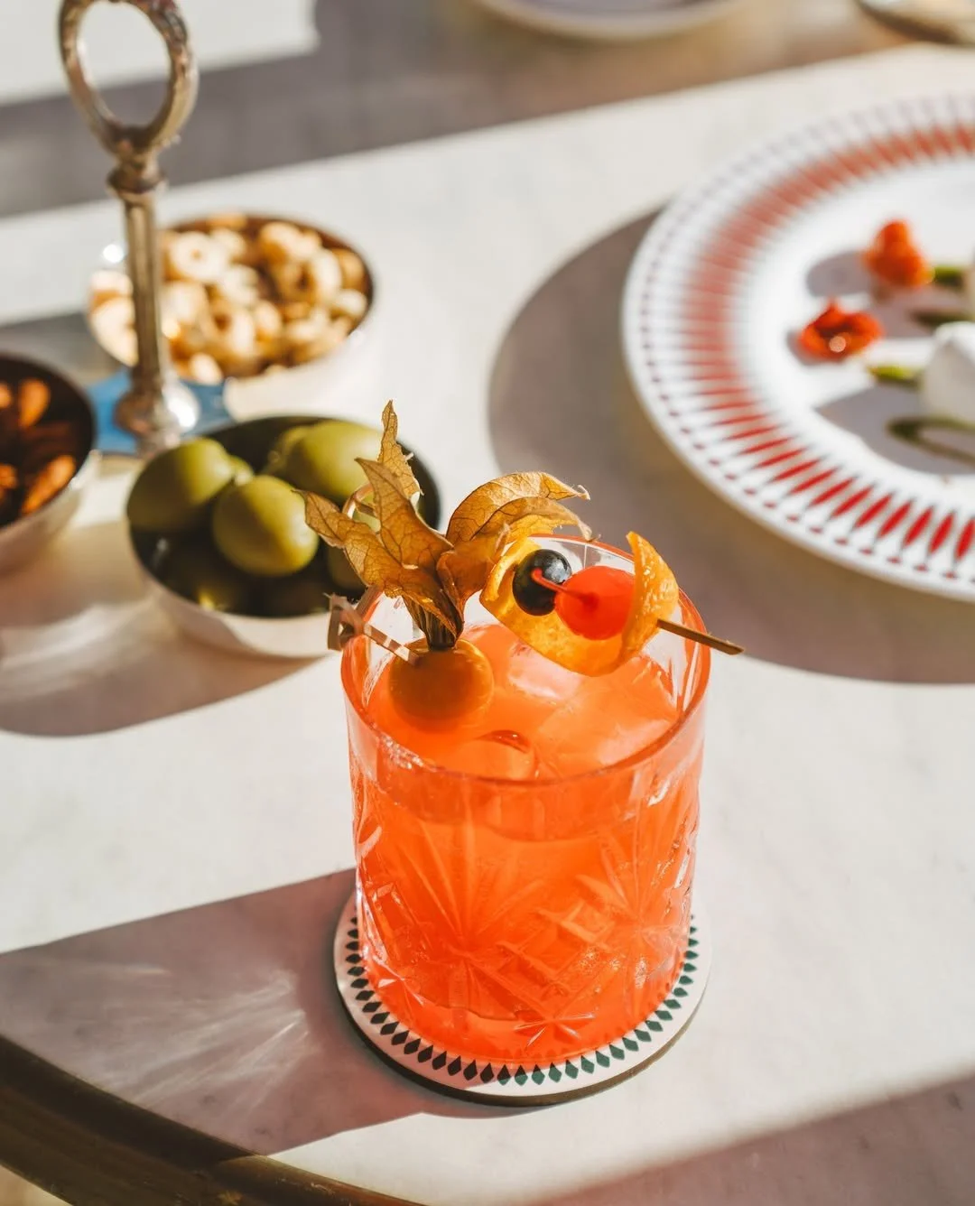 Colorful cocktail garnished with fruit and herbs on a white table. In the background, shelves with bowls of snacks and plates with food.
