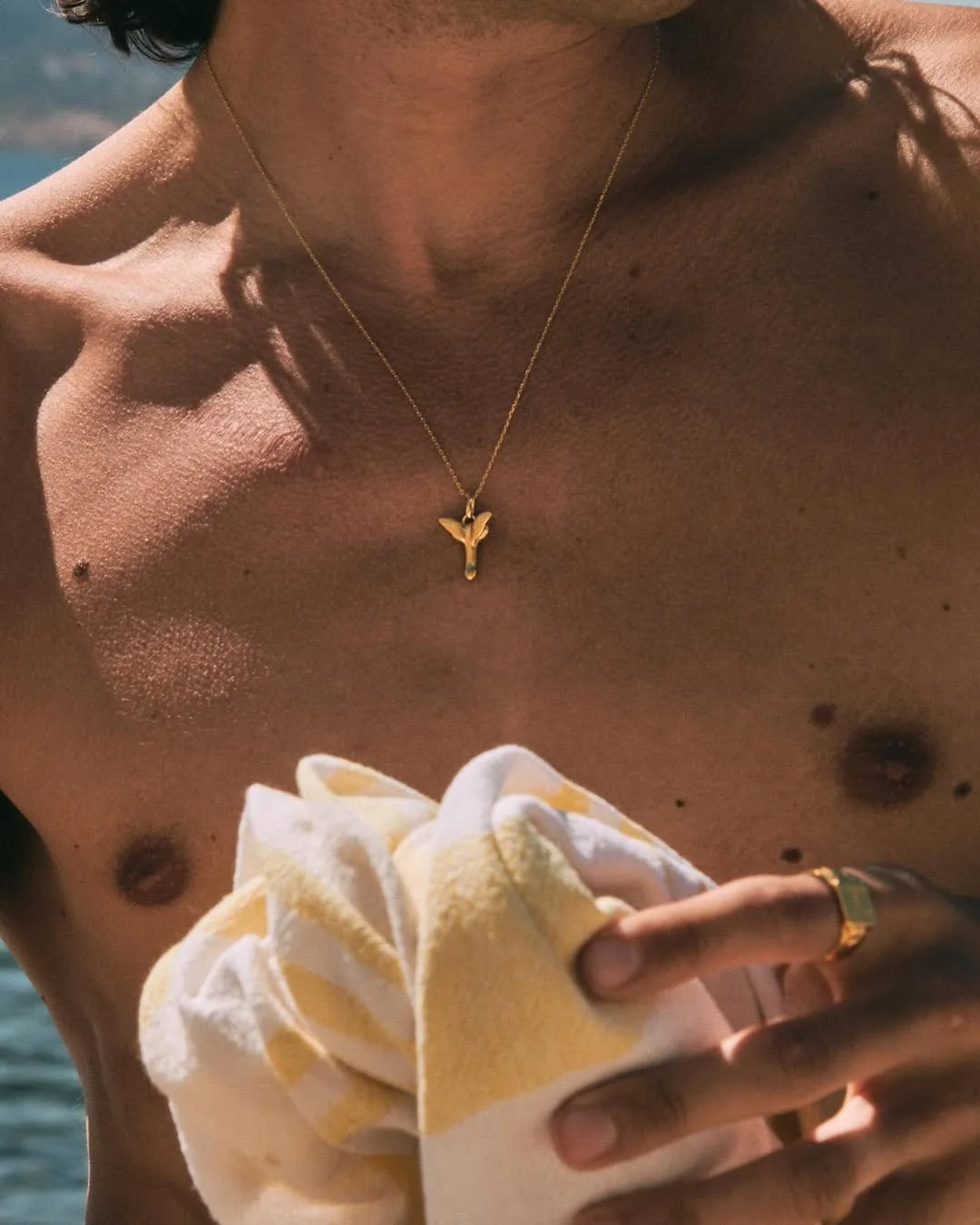 Close-up of a shirtless person wearing a gold necklace with a butterfly pendant, holding a yellow and white towel.