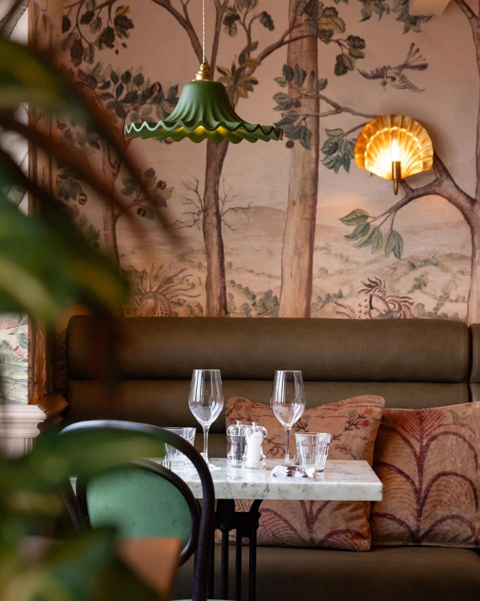 Interior of a restaurant with a marble table set with glasses and salt & pepper shakers, a green pendant light, a wall with a mural of trees and wildlife, and a cushioned banquette with patterned pillows.
