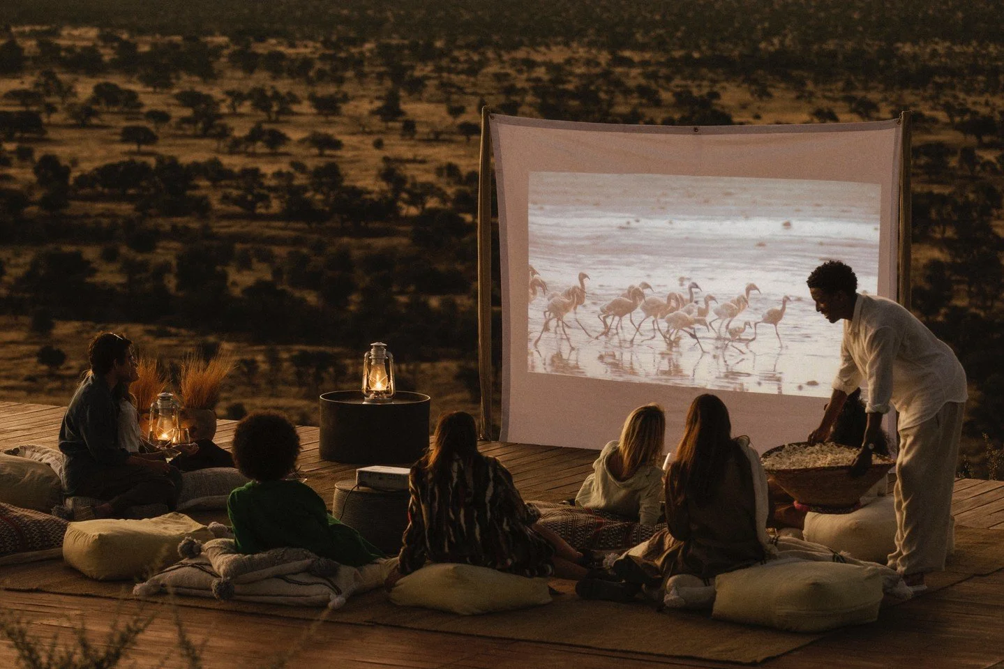 People watching a movie of flamingos on a large outdoor screen at night, with a person serving snacks, surrounded by lanterns and cushions on a wooden deck in a desert landscape.