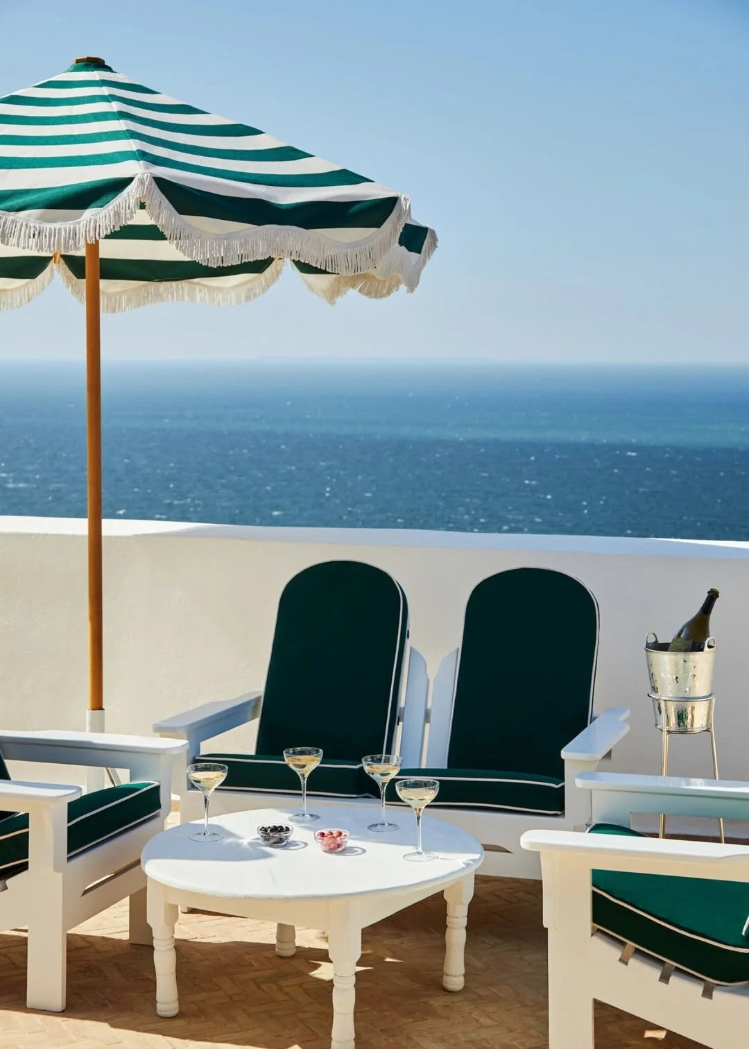 Ocean view from a terrace with a green and white striped umbrella, white furniture with green cushions, a small white table with drinks and snacks, and a bucket with a bottle, overlooking the sea under a clear blue sky.