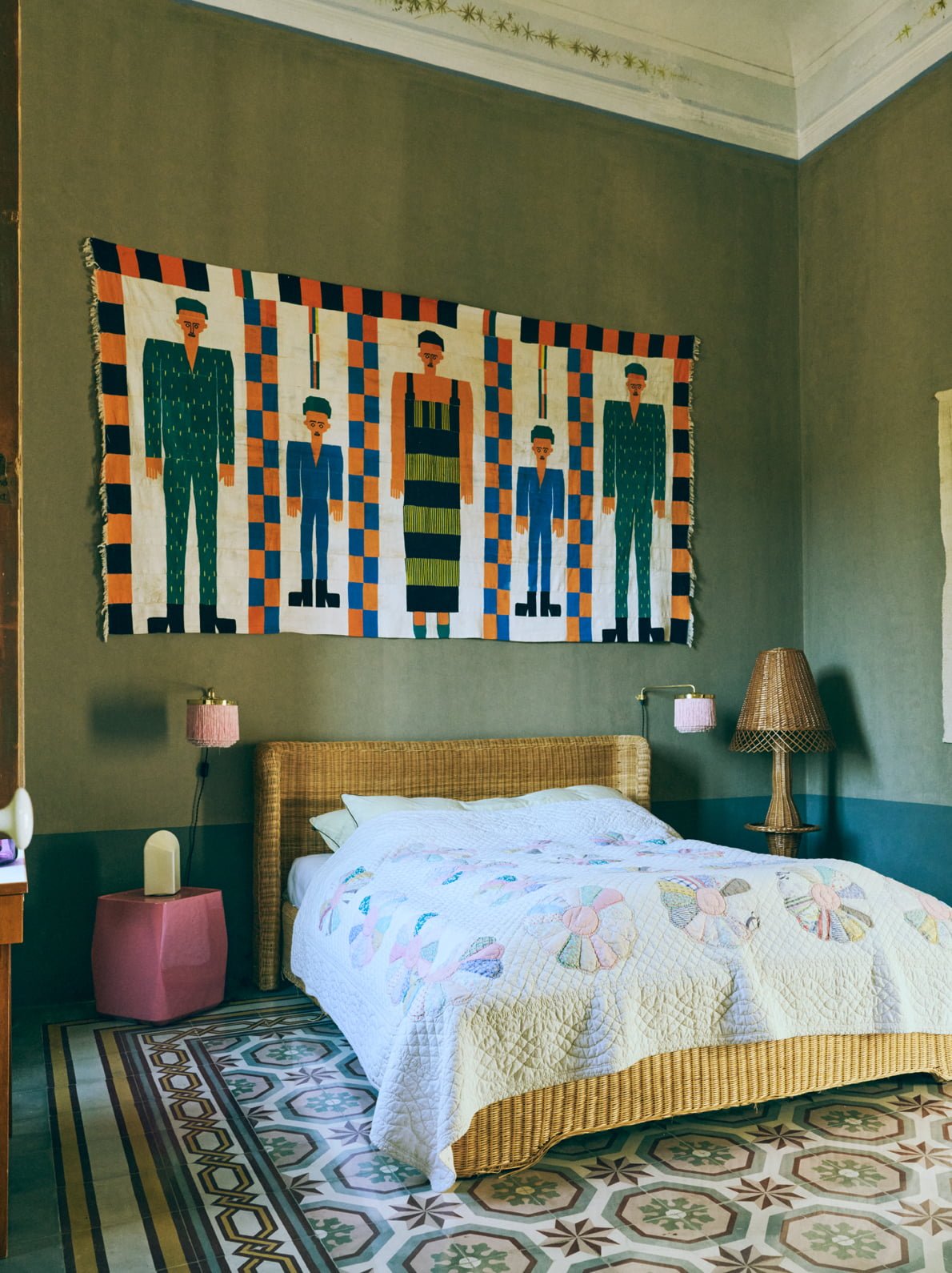 A bedroom with a wicker bed, colorful quilt, wall art depicting five stylized figures, pink lamps, a wicker table, patterned floor tiles, and green walls.