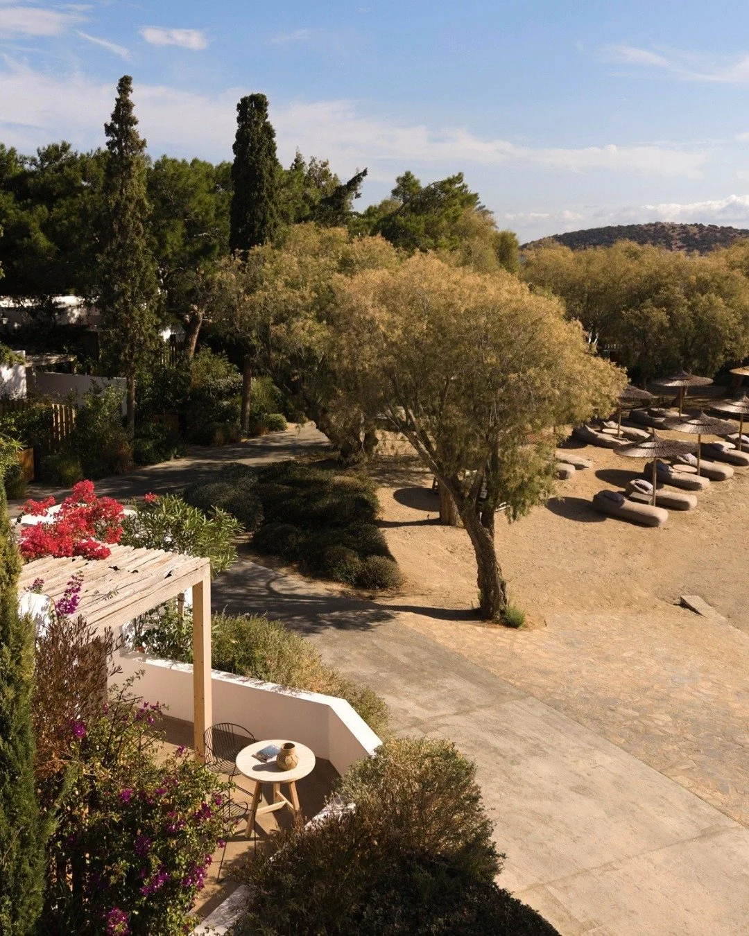 A peaceful outdoor scene featuring a sandy area with lounge chairs and umbrellas, surrounded by trees and a small patio with a table and chairs, with a blue sky and some clouds overhead.