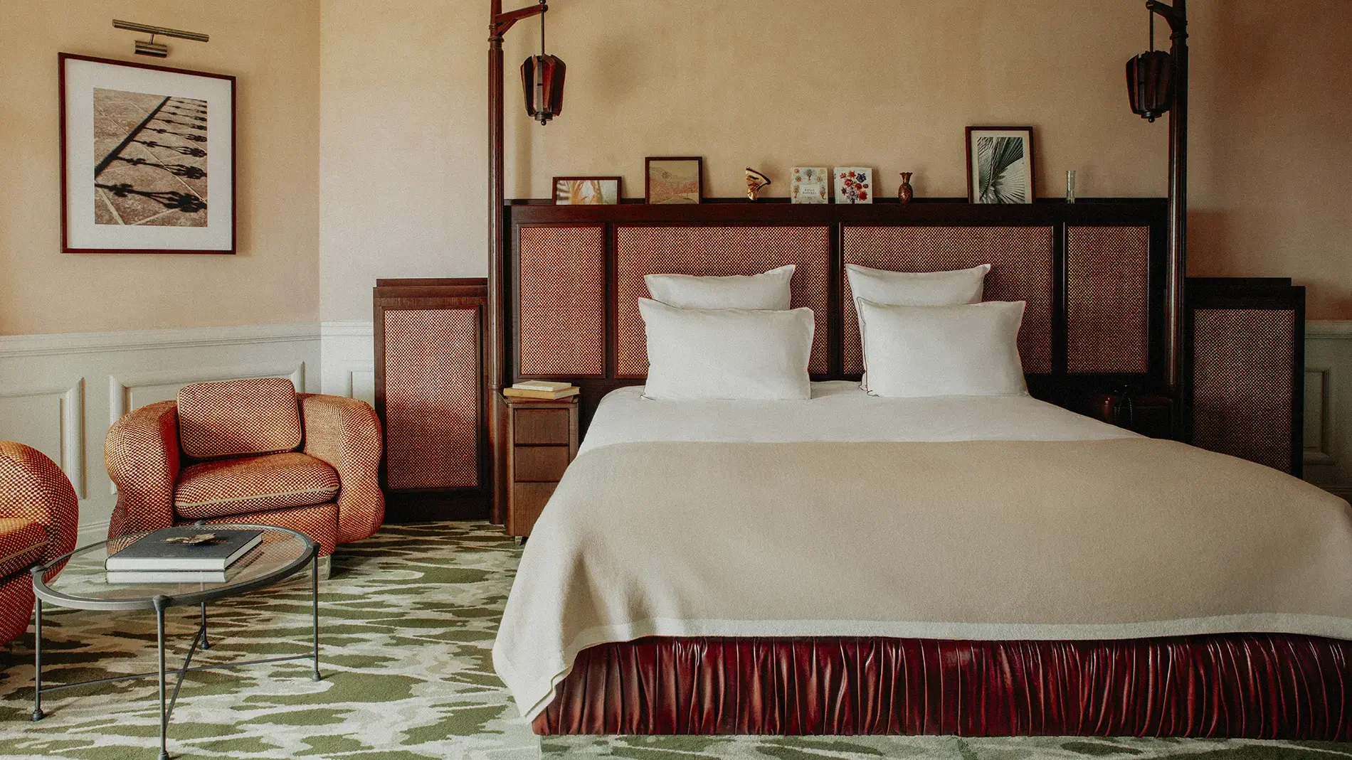 Hotel bedroom with a large bed, white pillows, a wooden headboard with decor, a side table with books, an orange armchair, a glass-top coffee table with magazines, framed artwork, and wall-mounted lamps.