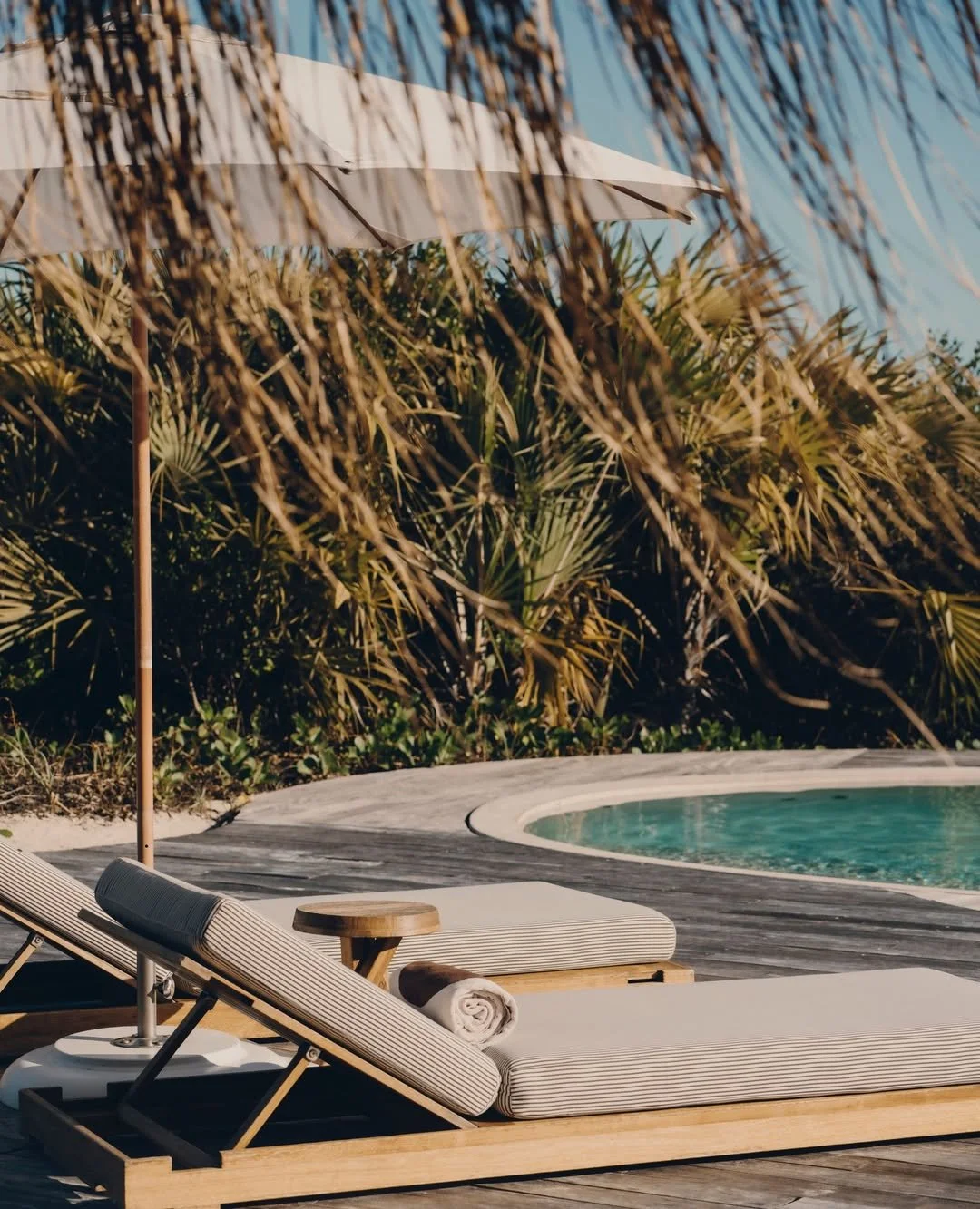 A poolside area with two beige lounge chairs, a rolled white towel on one, a small round wooden side table, and a white umbrella, surrounded by dense greenery and palm trees.