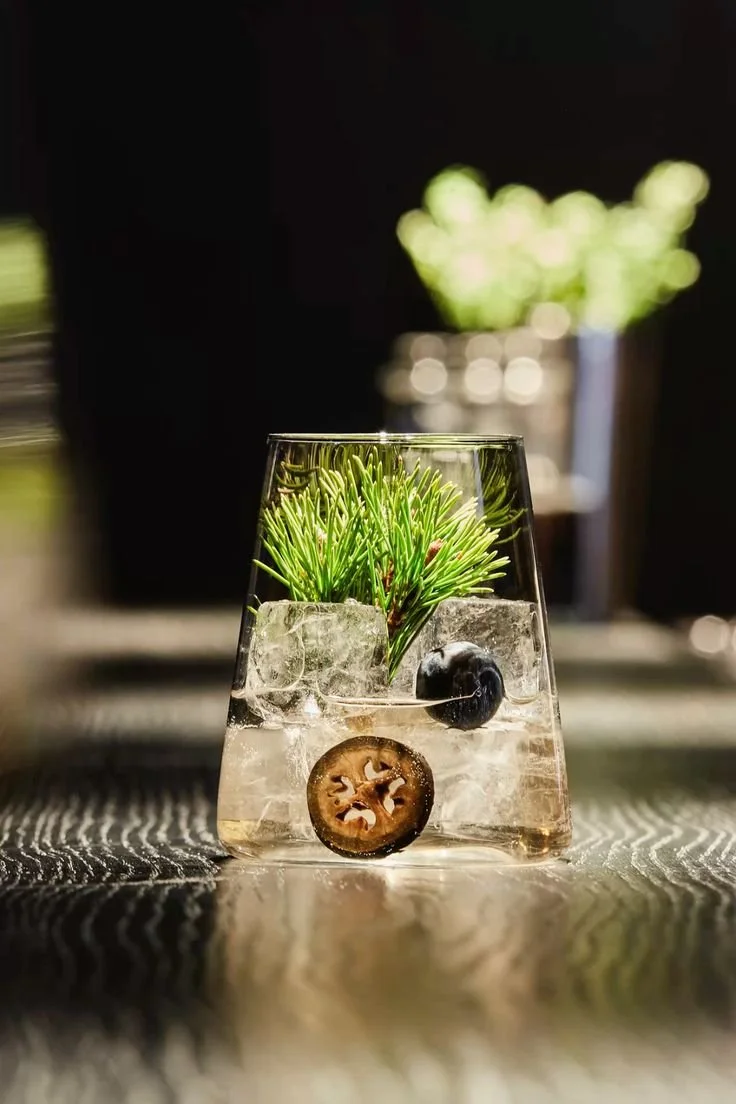 Glass with ice, blackberries, and fresh green pine branches, with another blurred plant in the background.