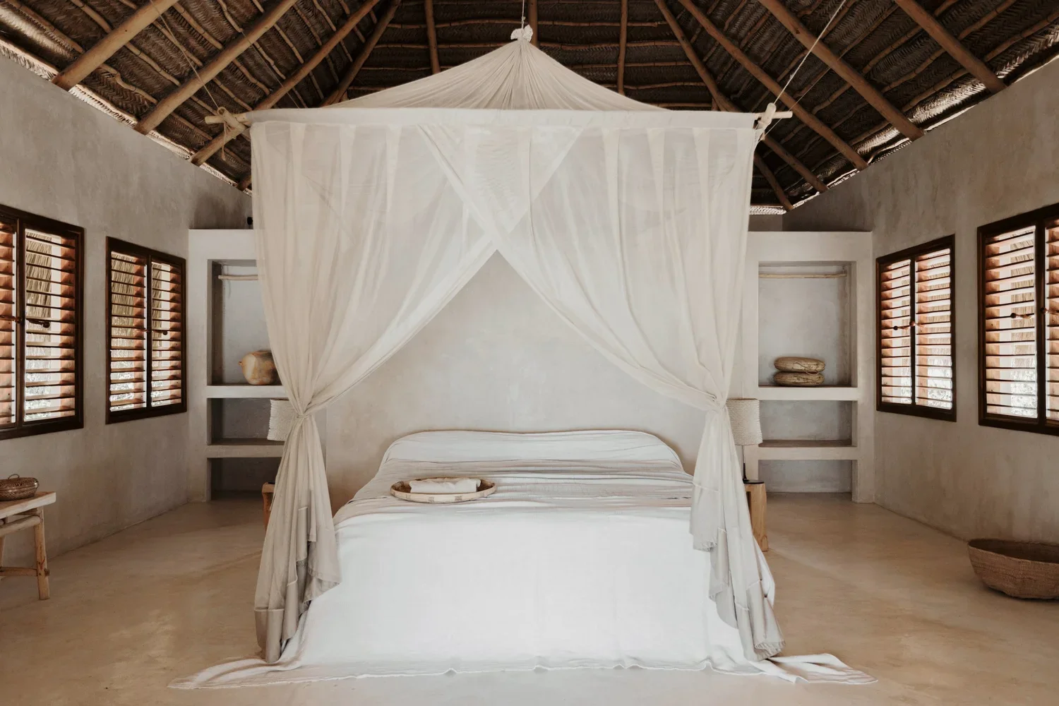 A white bed with a canopy in a rustic, minimalist bedroom with wooden window shutters, a thatched ceiling, and neutral decor.