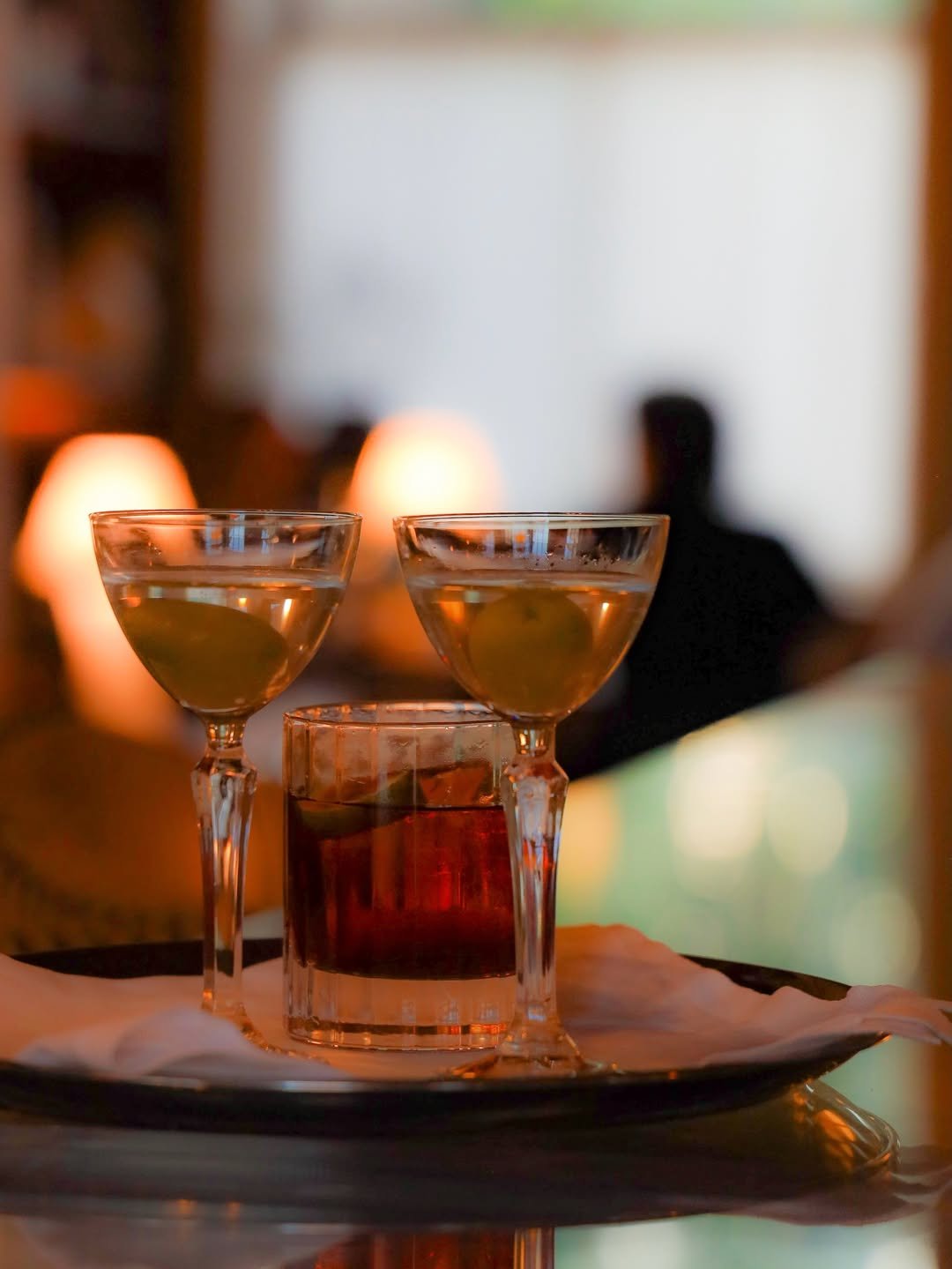 Two cocktail glasses with a golden drink on a tray, with a candle and blurred background of lamps and a person.