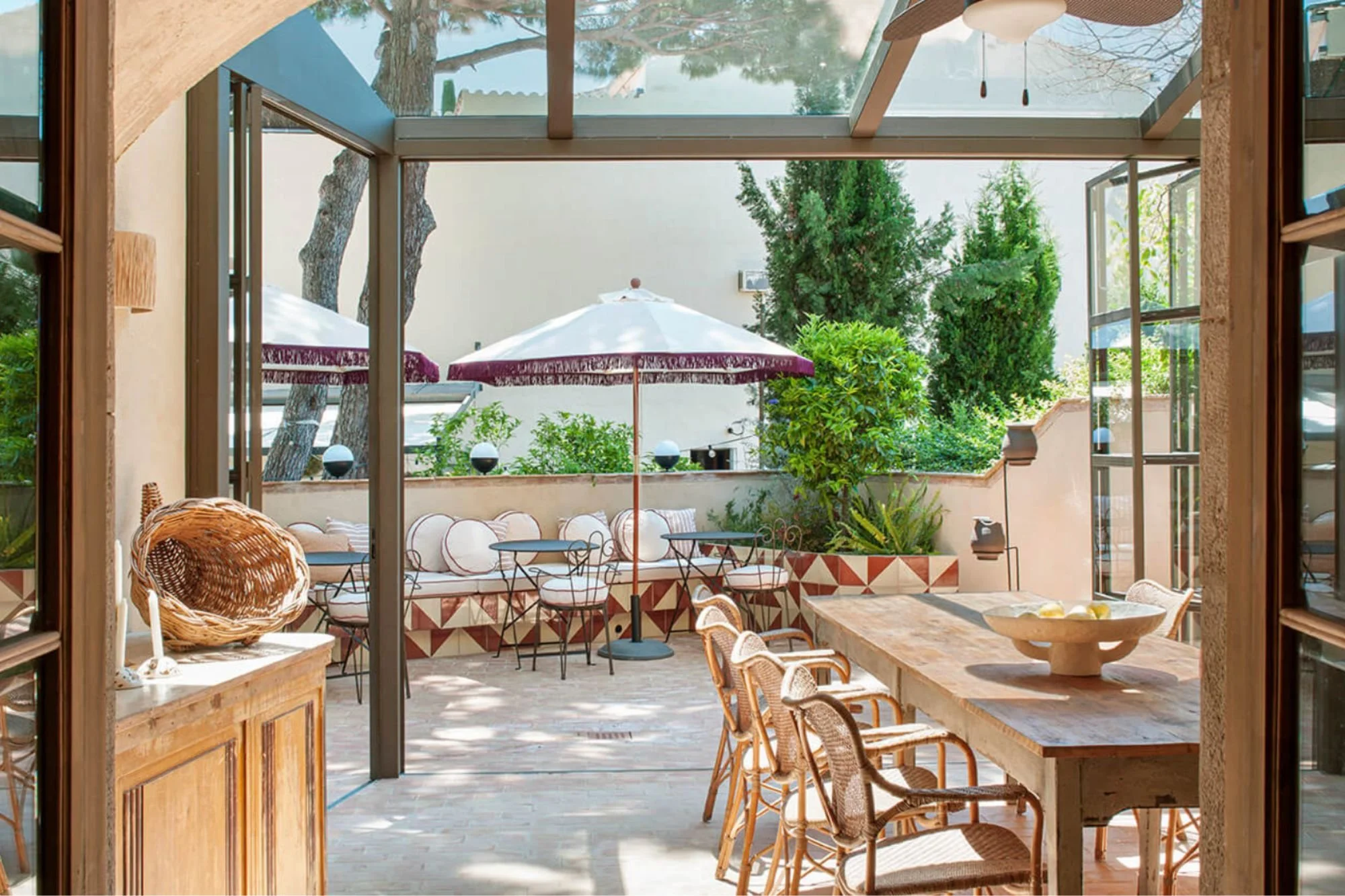 A cozy outdoor patio area with a large white patio umbrella, surrounded by lush greenery including trees and bushes, and furnished with wicker chairs and a low table, viewed through a glass sliding door.