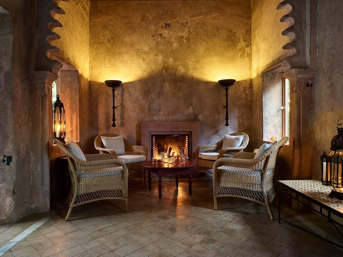 A cozy indoor seating area with four wicker chairs with white cushions arranged around a central small round wooden table in front of a fireplace with a lit fire. The walls are rustic with a warm yellowish-brown tone, and there are two wall-mounted sconces and two floor lamps providing soft lighting. The floor is tiled, and a window on the right side allows natural light to enter.