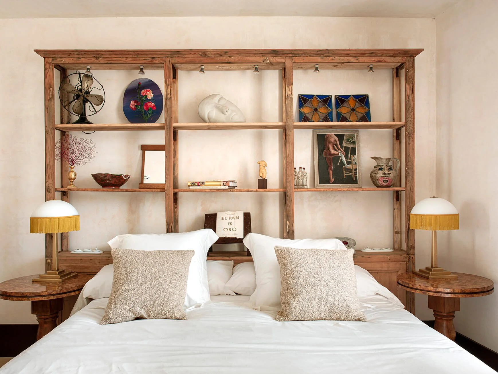 A bedroom with a large bed, white bedding, and beige pillows. Behind the bed is a wooden shelving unit with various decorative items, including a vintage fan, painted plates, a white sculpture, framed artwork, a vase, and books. Two matching lamps with yellow fringed shades are on round bedside tables on either side of the bed.