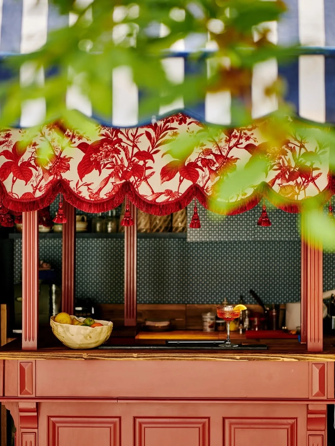 Pink bar with red and pink floral canopy, bowl of lemons and limes, cocktail glass with a drink and lemon wedge, seen through green leaves.