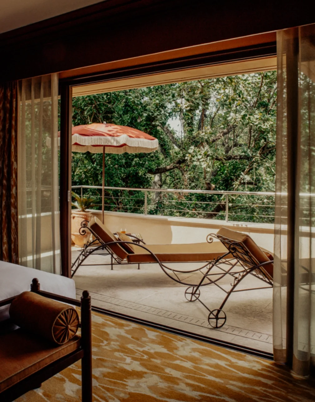 View of a balcony with lounge chairs, a large orange and white umbrella, and green trees in the background, seen through open glass sliding doors from inside a room.