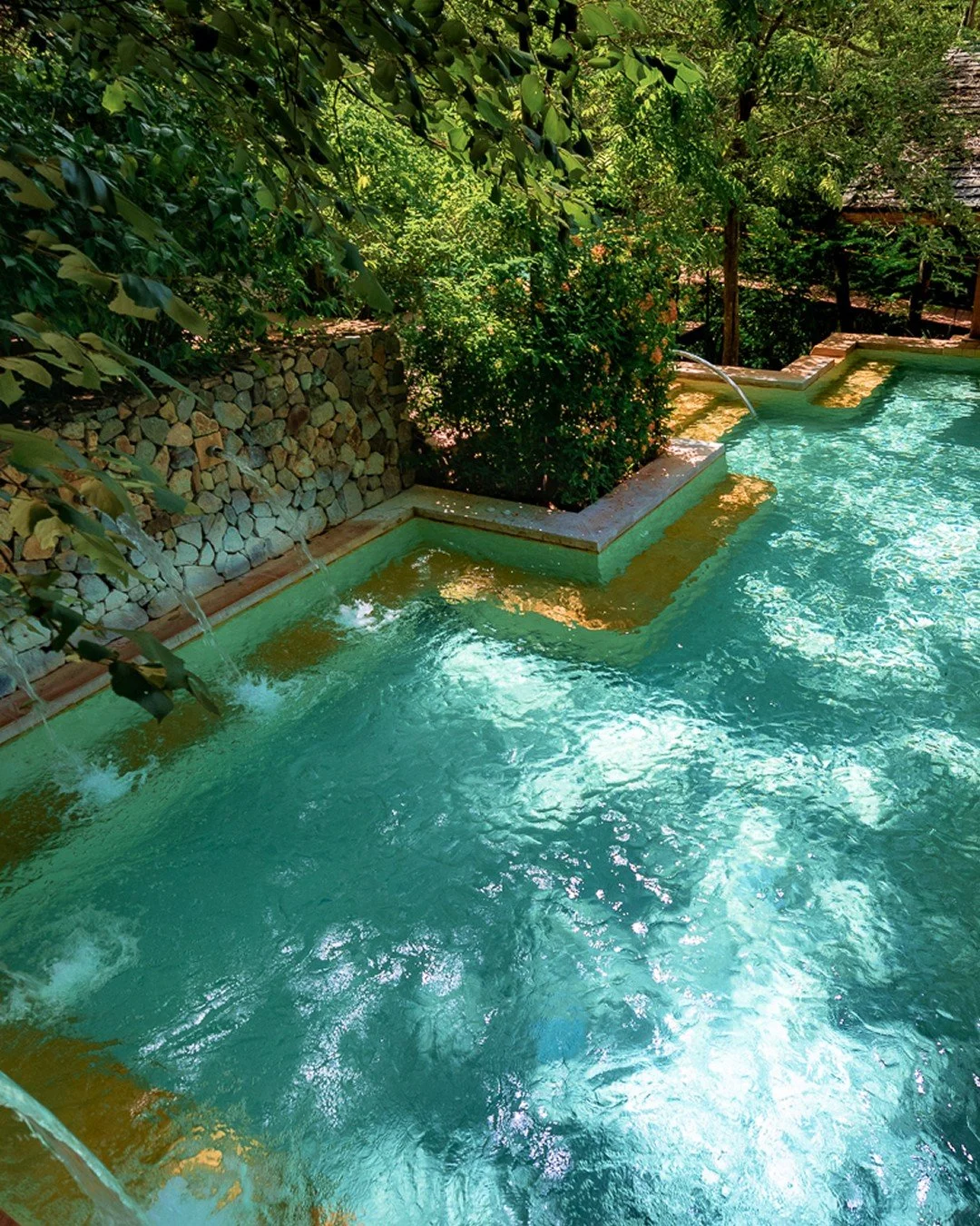 A scenic outdoor swimming pool with stone water features, surrounded by dense green foliage and trees, creating a tranquil and private setting.