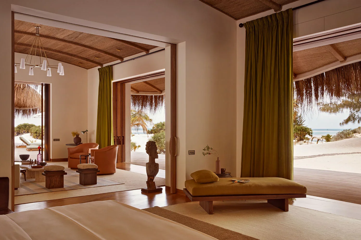 Luxurious beachside resort room with view of sandy beach, tropical trees, and ocean, featuring a sitting area with chairs, ottomans, and decorative items, and large open doors allowing natural light and fresh air.