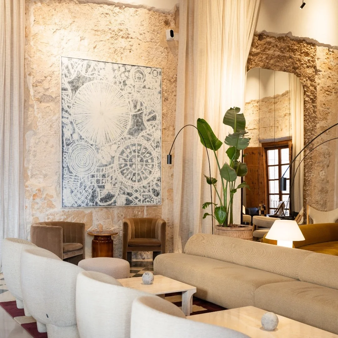 A stylish living room with exposed stone walls, beige curtains, modern artwork, large green plant, beige and brown furniture, and warm lighting.