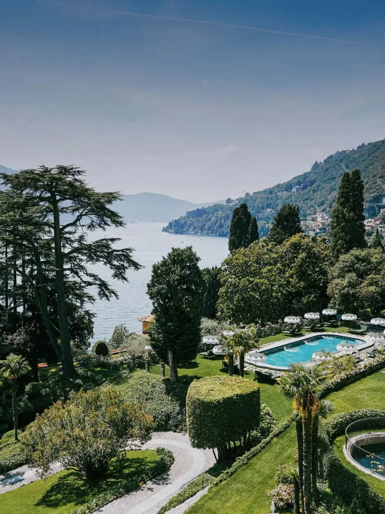 A lakeside resort with a swimming pool surrounded by lush greenery, tall trees, and a view of a calm lake with hills in the background.
