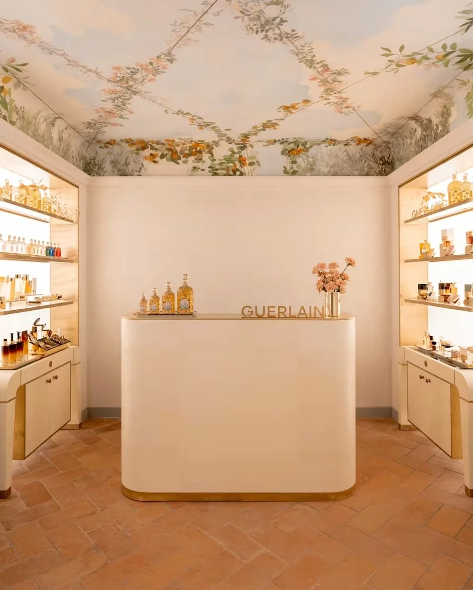 Interior of a Guerlain perfume boutique with white display shelves on either side, a curved central counter with the Guerlain logo, and floral decor including pink flowers and a painted ceiling with a floral pattern.