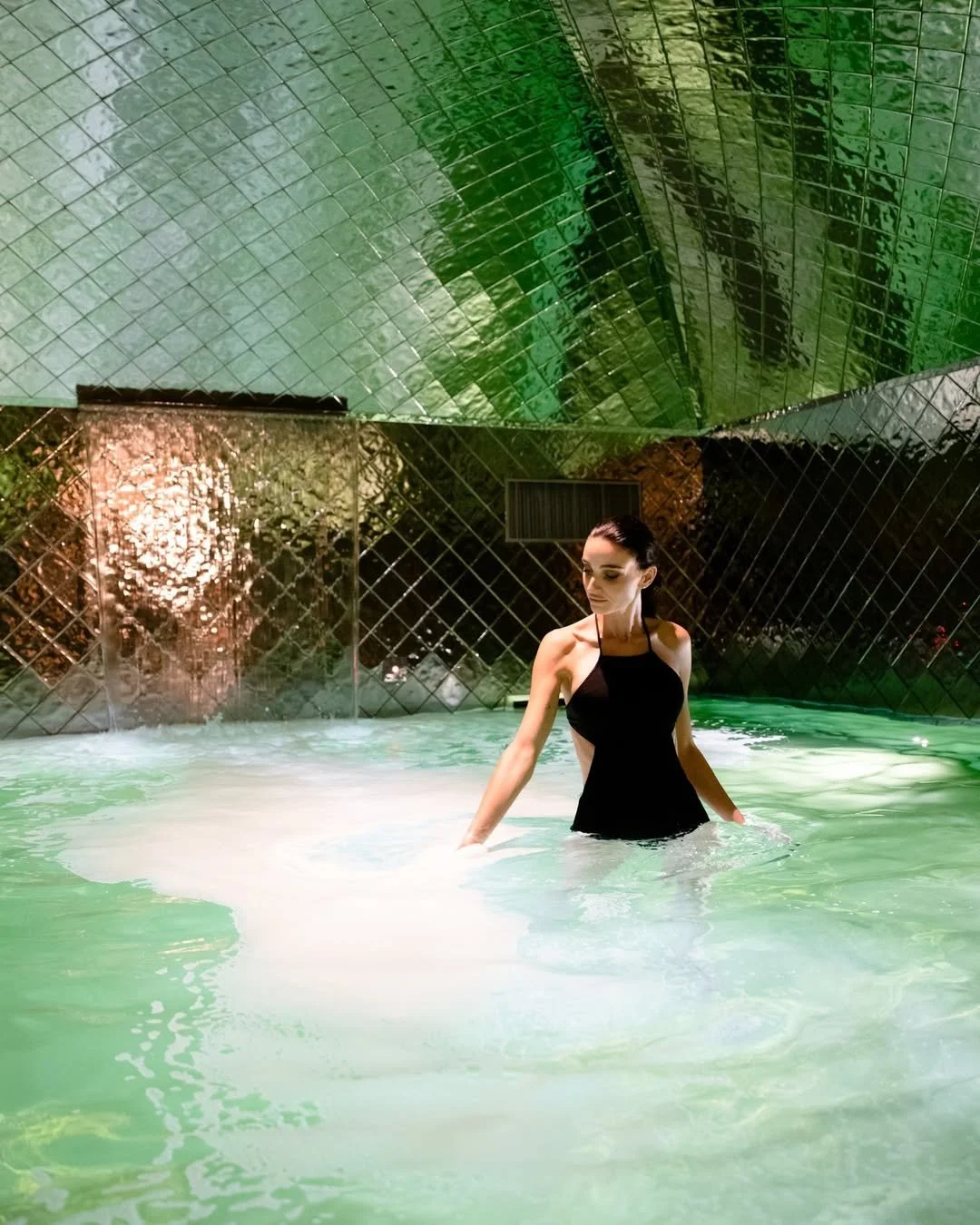 A woman in a black dress stands in a pool of water with a reflective green ceiling and a tiled wall background.