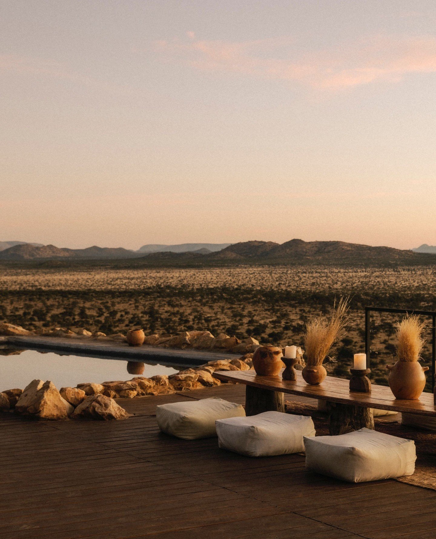 Outdoor patio with cushions, vases, candles, and a pool overlooking a desert with mountains at sunset.