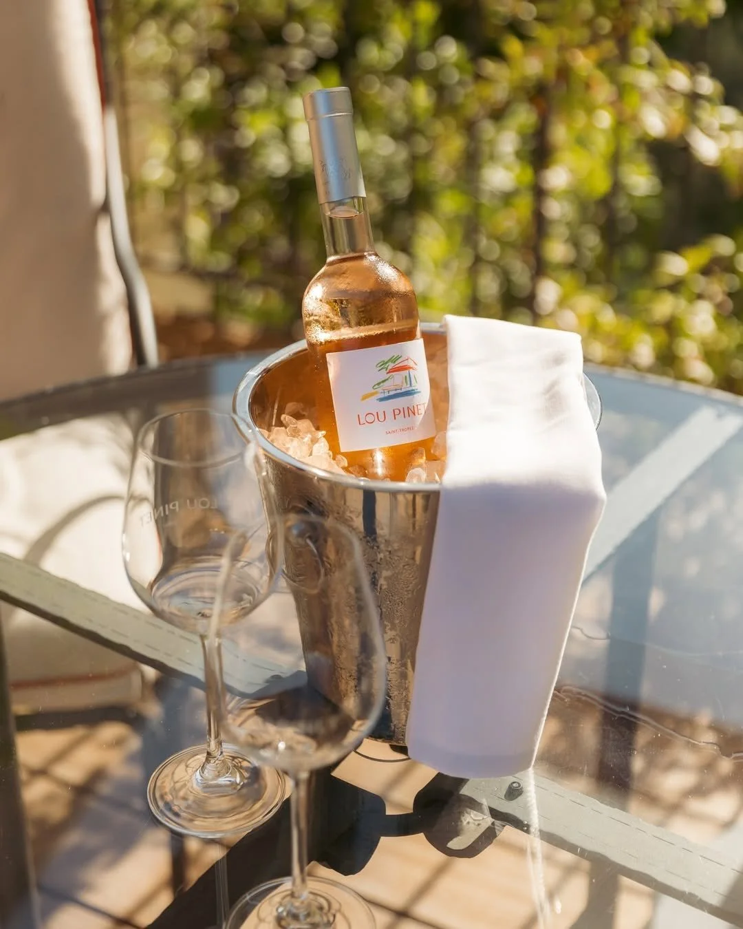 A bottle of rosé wine in an ice bucket with a white cloth, two empty wine glasses, and a small empty glass on a glass table outdoors with green and yellow foliage in the background.