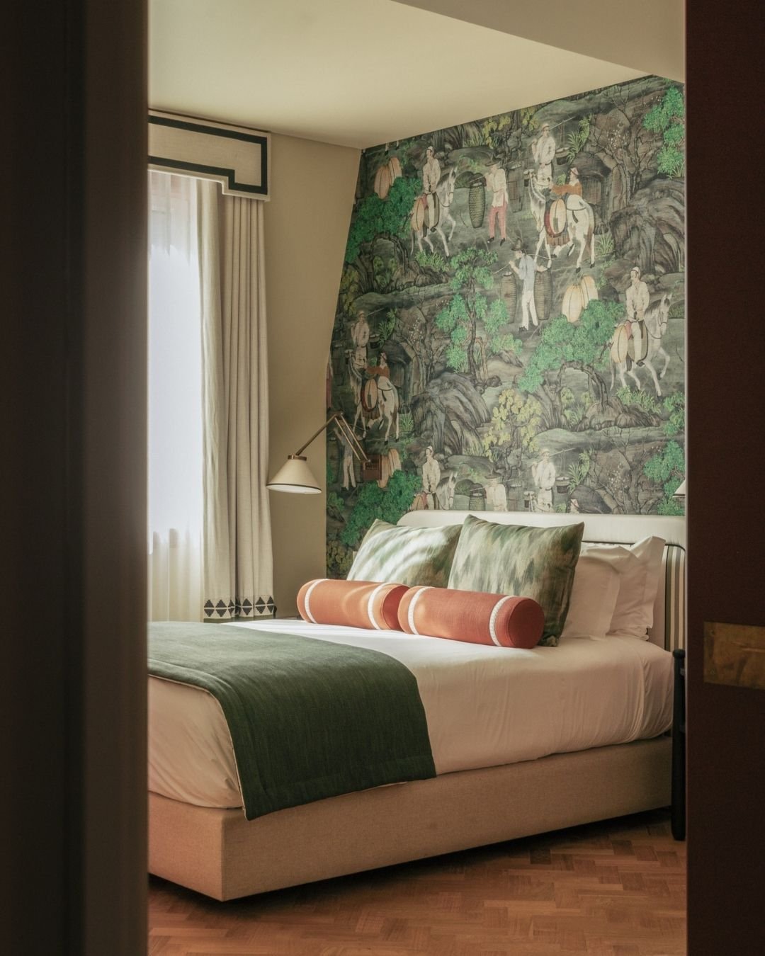 A cozy hotel bedroom with a bed, green and orange pillows, a green bed runner, and a decorative mural on the wall featuring traditional Asian figures and nature scenes. The room has a window with white curtains and a wall-mounted reading lamp.