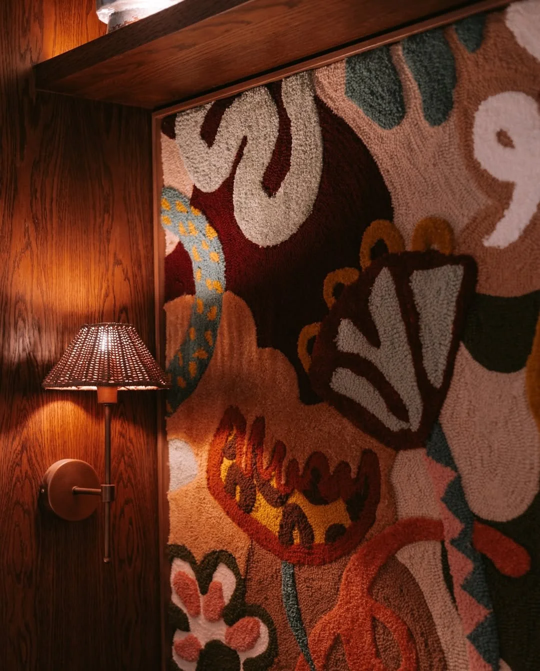 A wall-mounted lamp with a woven shade illuminates a colorful floral-patterned rug on a wooden wall.