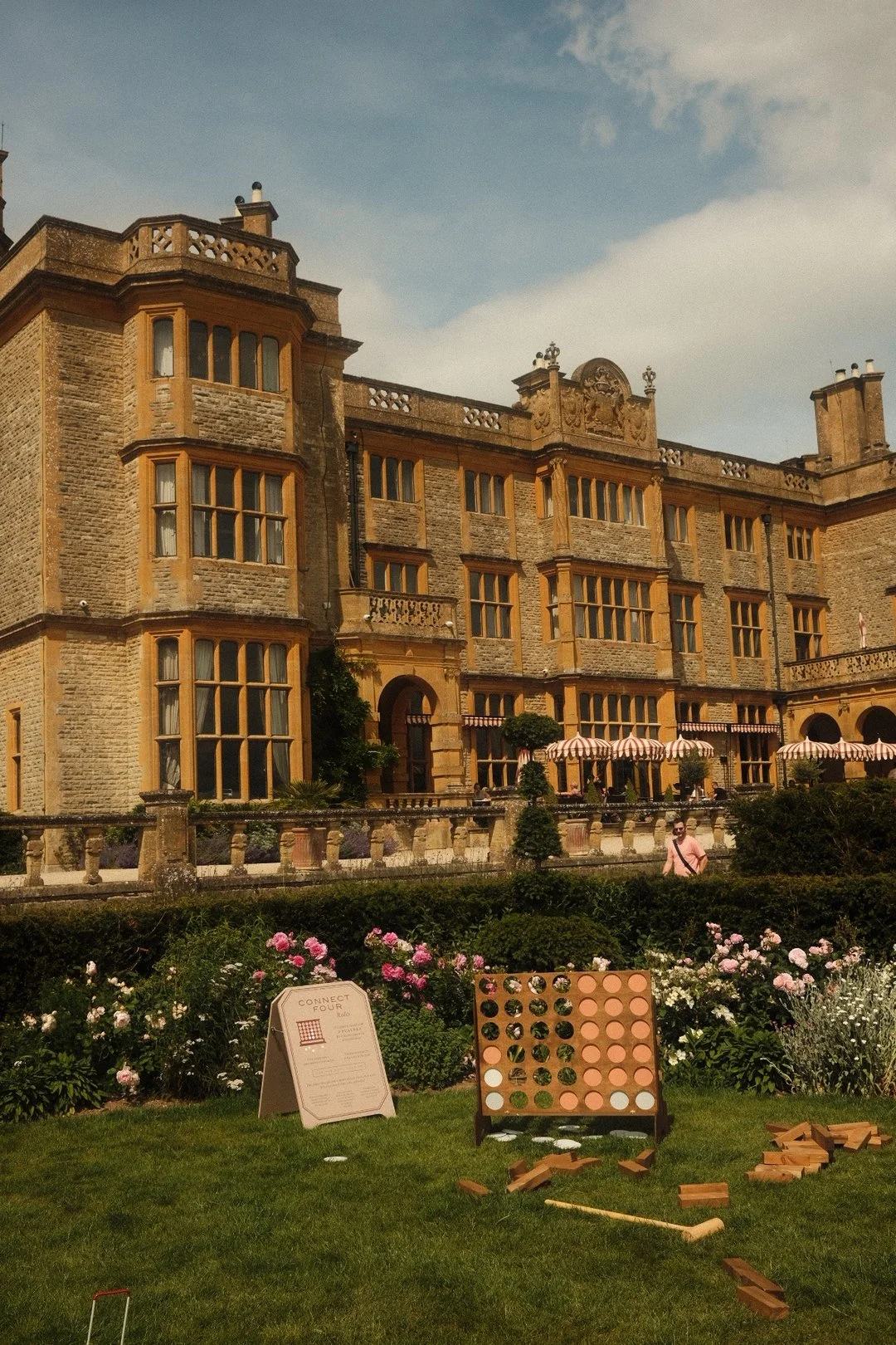 A large historic building with multiple stories, bay windows, and decorative details, set behind a garden with pink and white flowers. In the garden, there is a large Connect Four game, pieces, and a sign with rules.