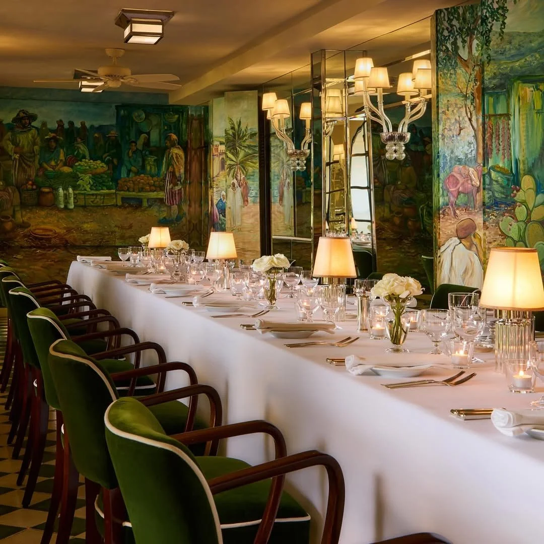 Elegant dining room with a long white table set with white napkins, glasses, candles, and flower arrangements, surrounded by green-upholstered chairs with wooden arms, and illuminated by warm table lamps and chandeliers. The background features colorful murals depicting outdoor scenes and people.