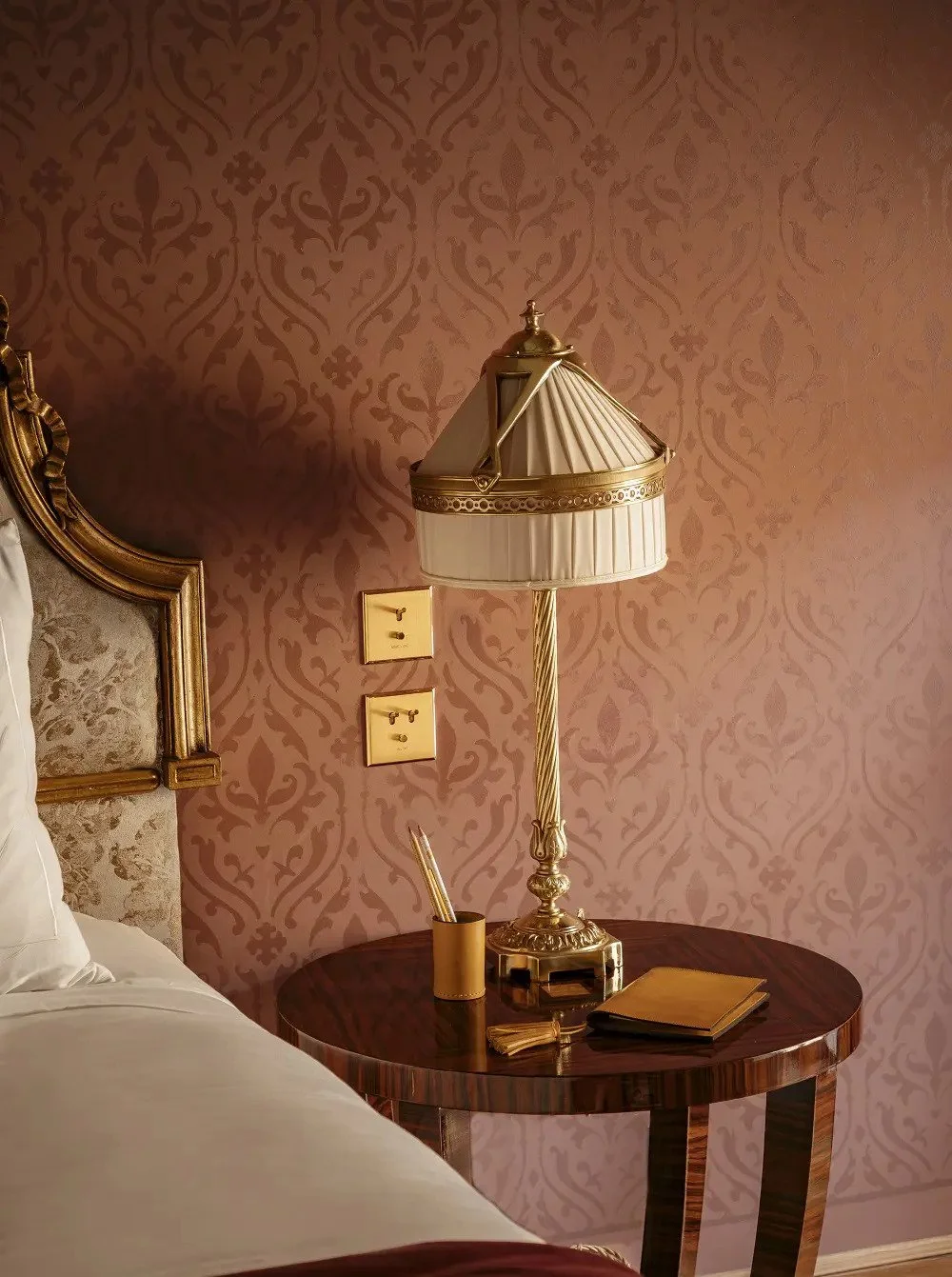 A classic interior bedroom scene with a wooden bedside table, a vintage lampshade with gold accents, a small notebook, and writing utensils. The wall has ornate pink damask wallpaper, and part of a bed with a gold-framed headboard is visible.